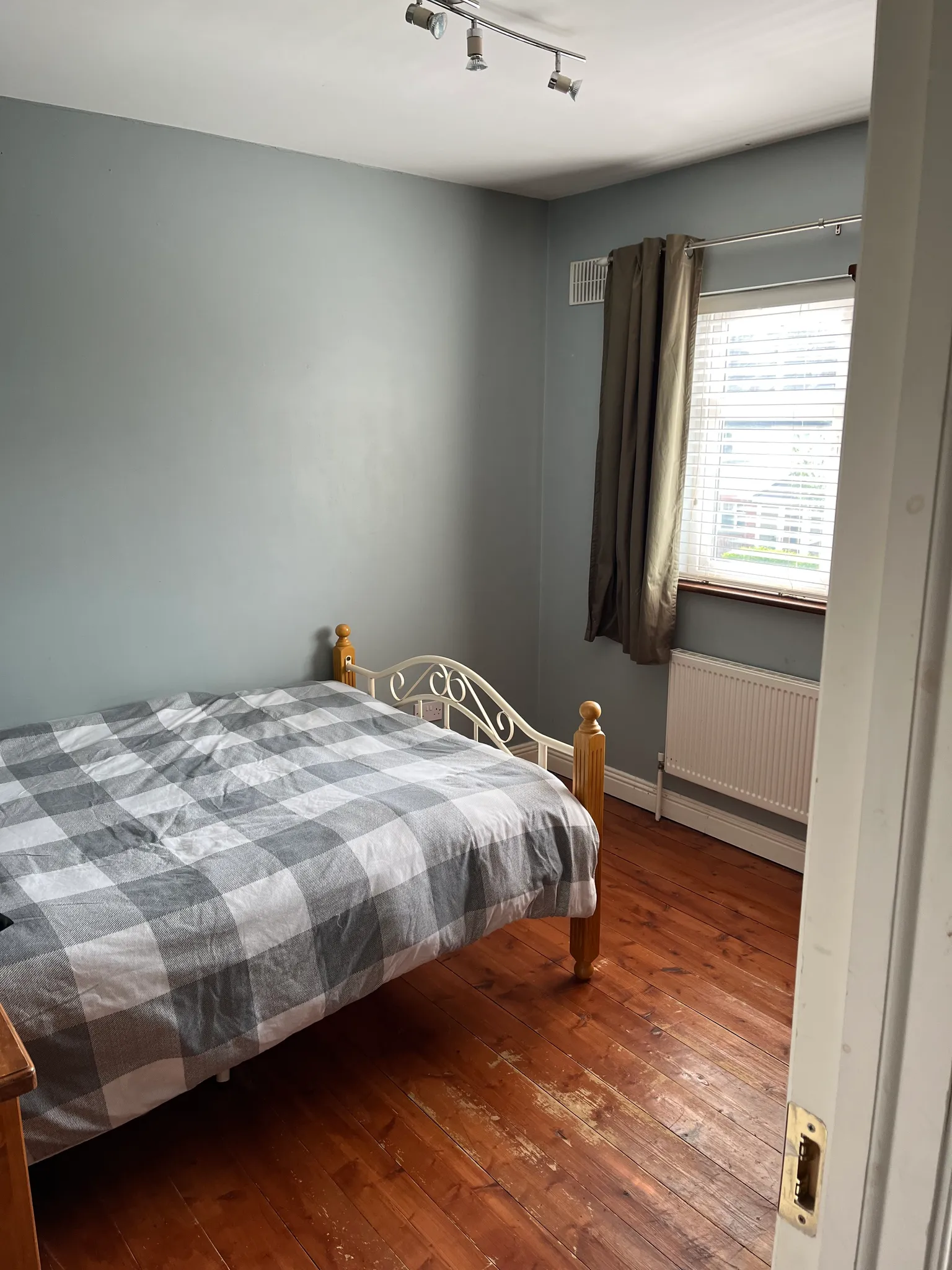Bedroom with a double bed with gray and white checkered bedding, metal bed frame with wooden finials, hardwood flooring, gray walls, window with brown and cream curtains, radiator, track lighting, and wooden door.