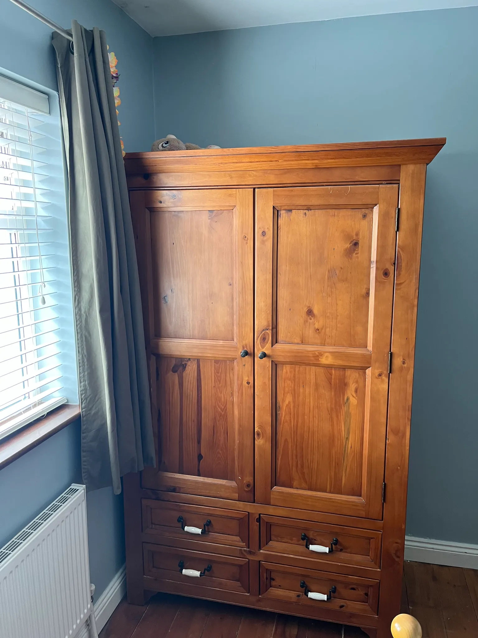 Bedroom with blue walls, wooden wardrobe with two doors and four drawers, window with blinds and gray curtains, and radiator.