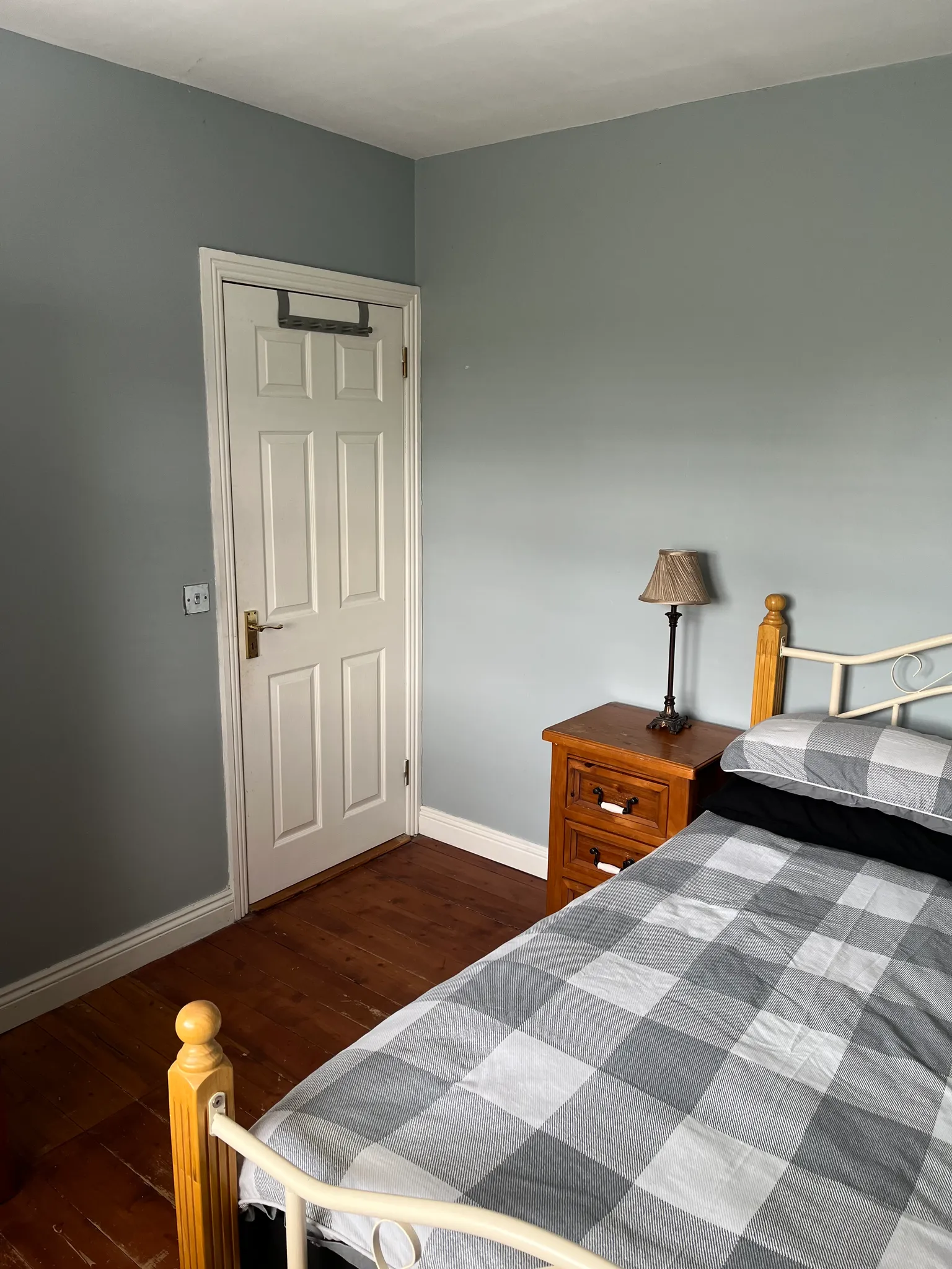 Bedroom with two single beds with gray and white checkered bedding, wooden nightstand with lamp, wooden bed frames, gray walls, white ceiling, dark wood flooring, and white six-panel door with black transom window.