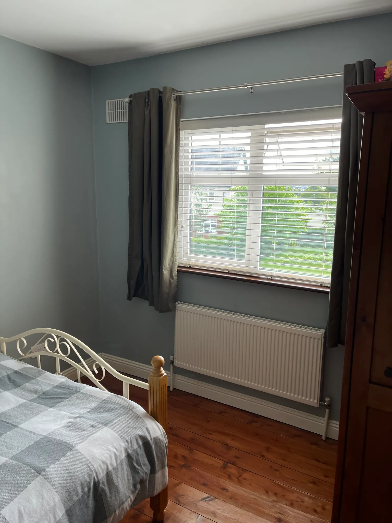 Bedroom with a single bed with metal headboard and checkered bedding, large window with horizontal blinds and dark curtains, radiator, wooden flooring, and gray-blue walls.