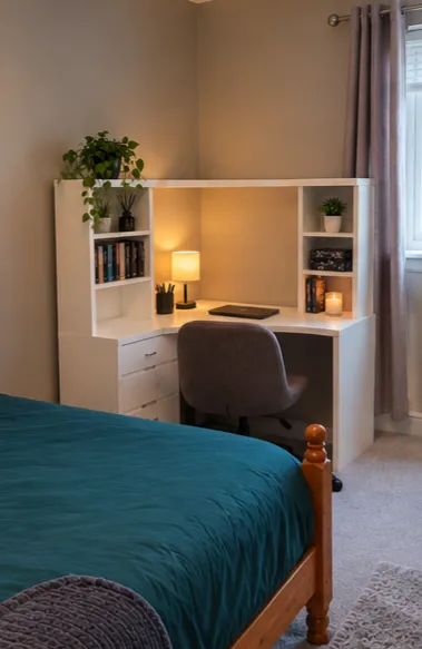 Bedroom with a wooden bed and teal bedcover, a white desk with hutch, swivel chair, table lamp, books and potted plants, and a window with curtains.