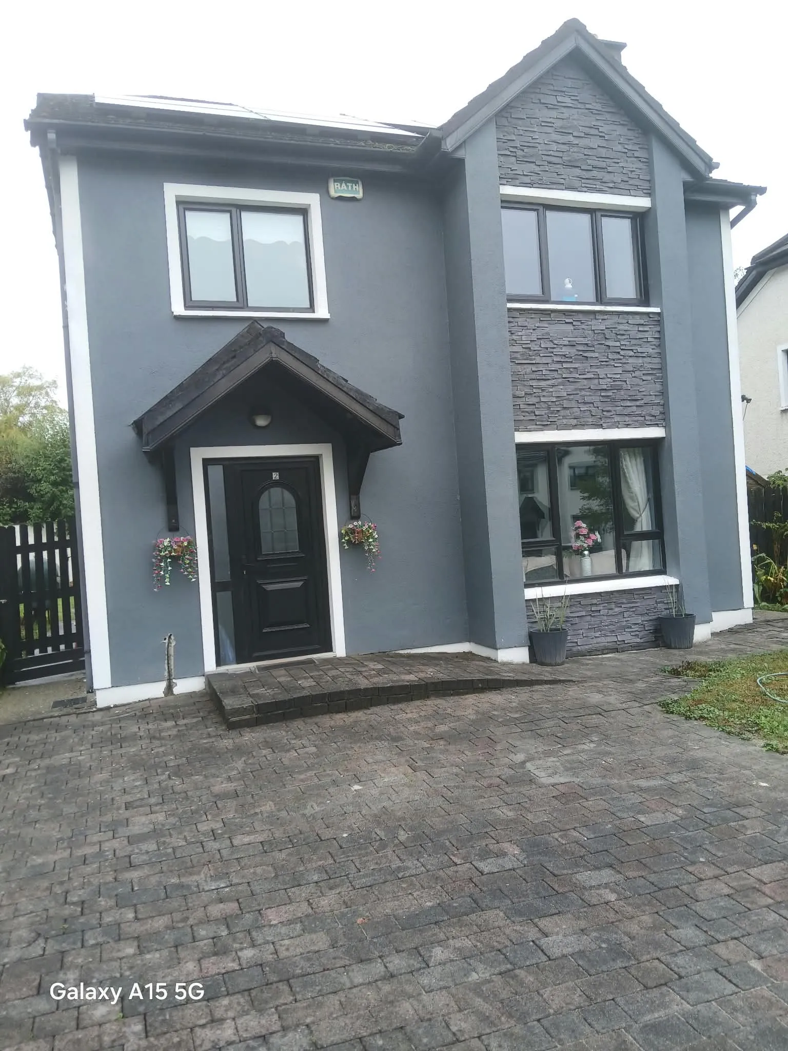 Front exterior of a two-storey grey house showing a black front door with porch, multiple windows, stone cladding on a bay section, potted plants and a paved driveway.