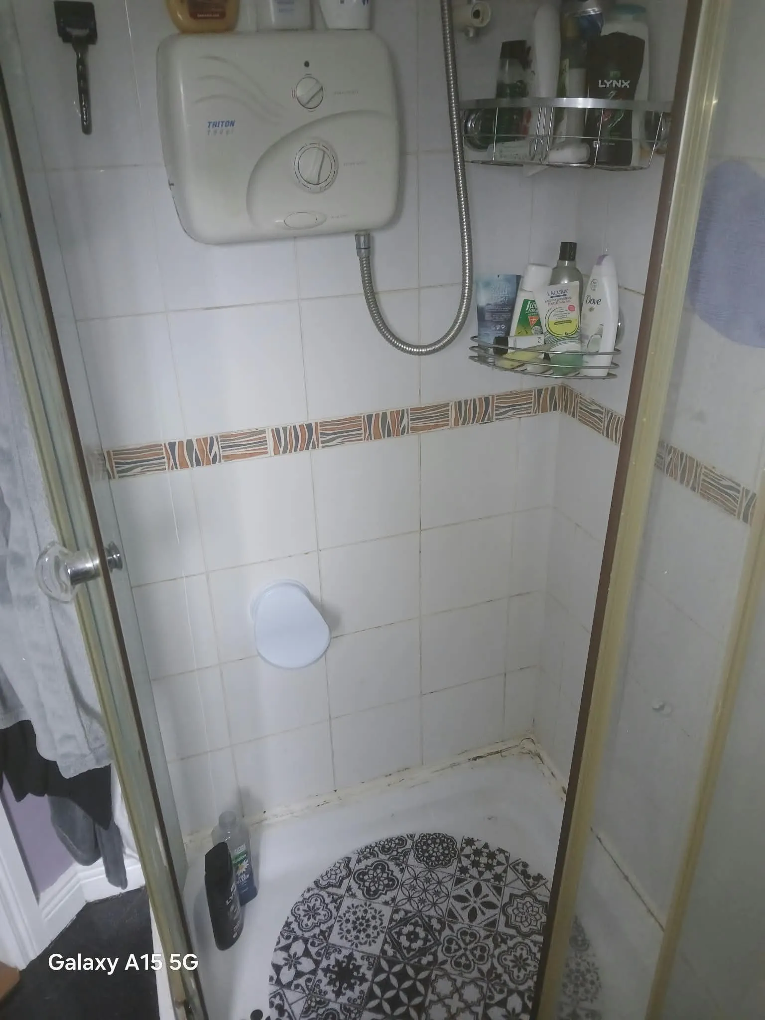 Tiled shower stall with a wall-mounted electric shower unit, flexible shower hose, corner metal shelf holding toiletries, and a patterned round shower mat.