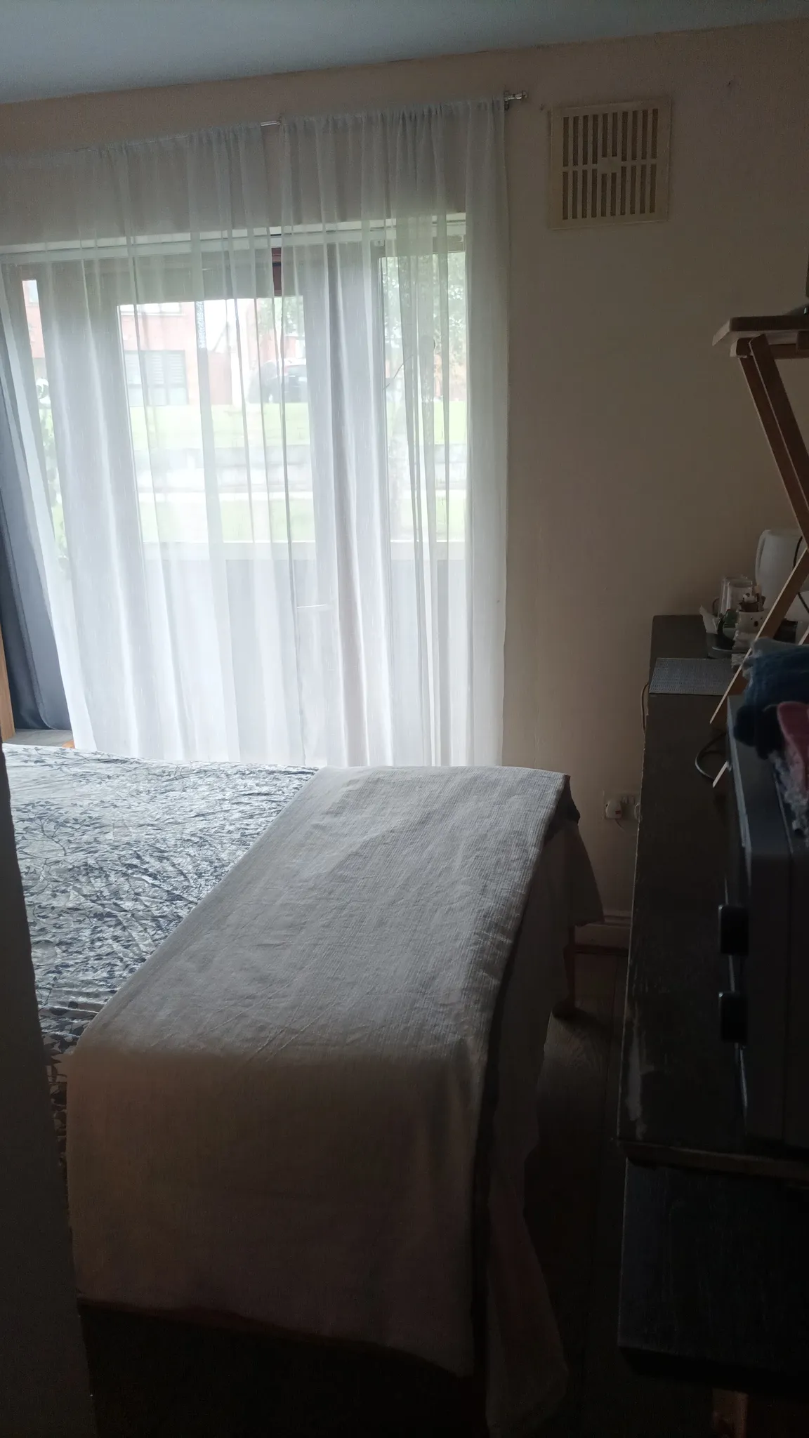 Bedroom showing a bed with a fabric-covered bench at the foot, sheer curtains covering a large window or sliding door and a dresser along the right wall.