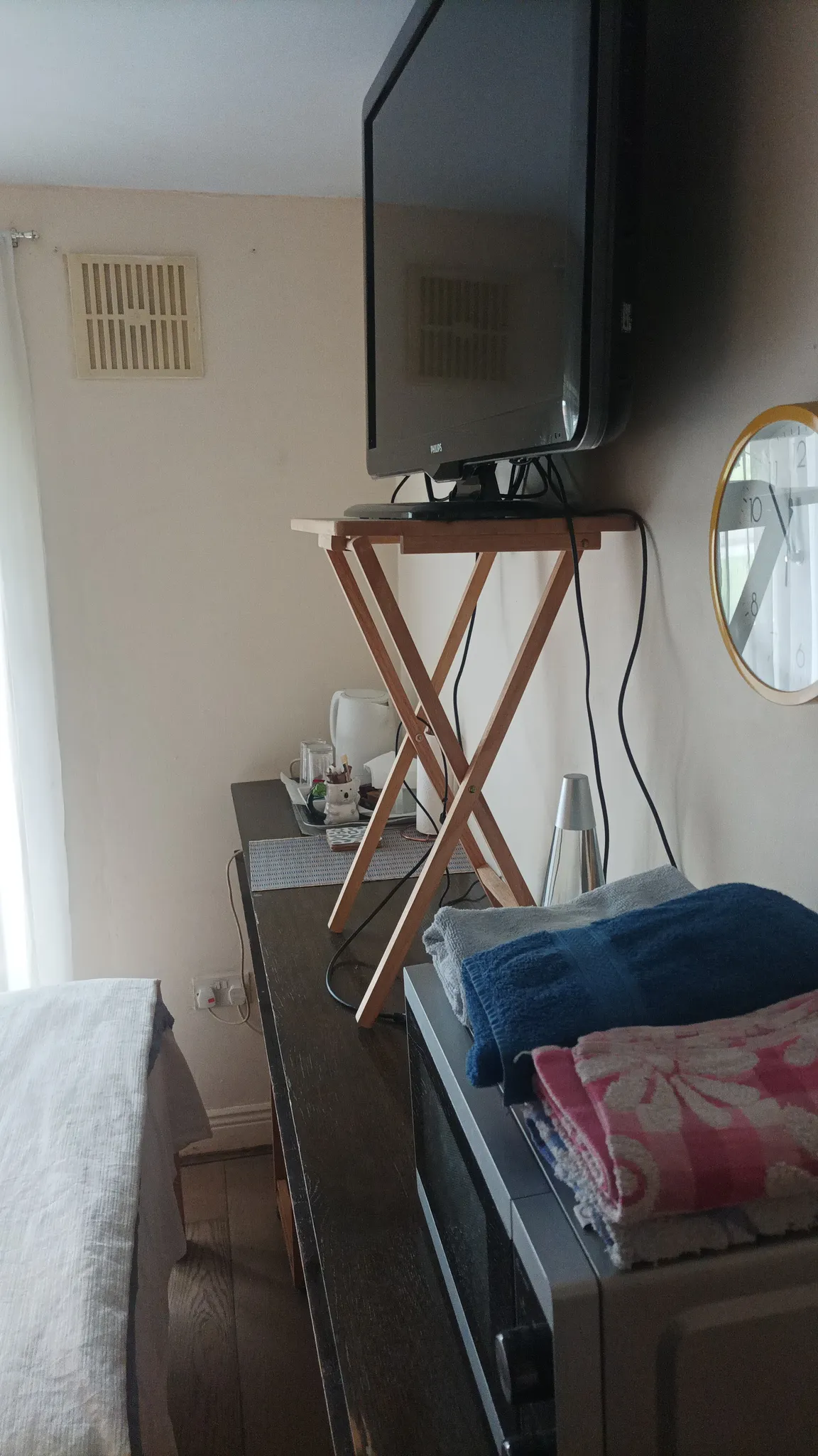 Bedroom area with a television on a small folding tray table placed on a long dresser, folded towels on a nearby appliance, a kettle and tray on the dresser, and a wall clock.