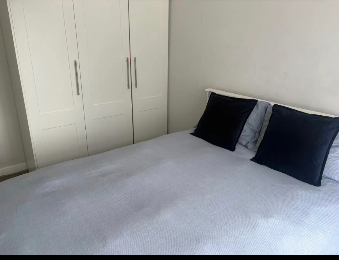 Bedroom with a double bed covered by a light-colored duvet, two dark pillows and a white built-in wardrobe along the wall.