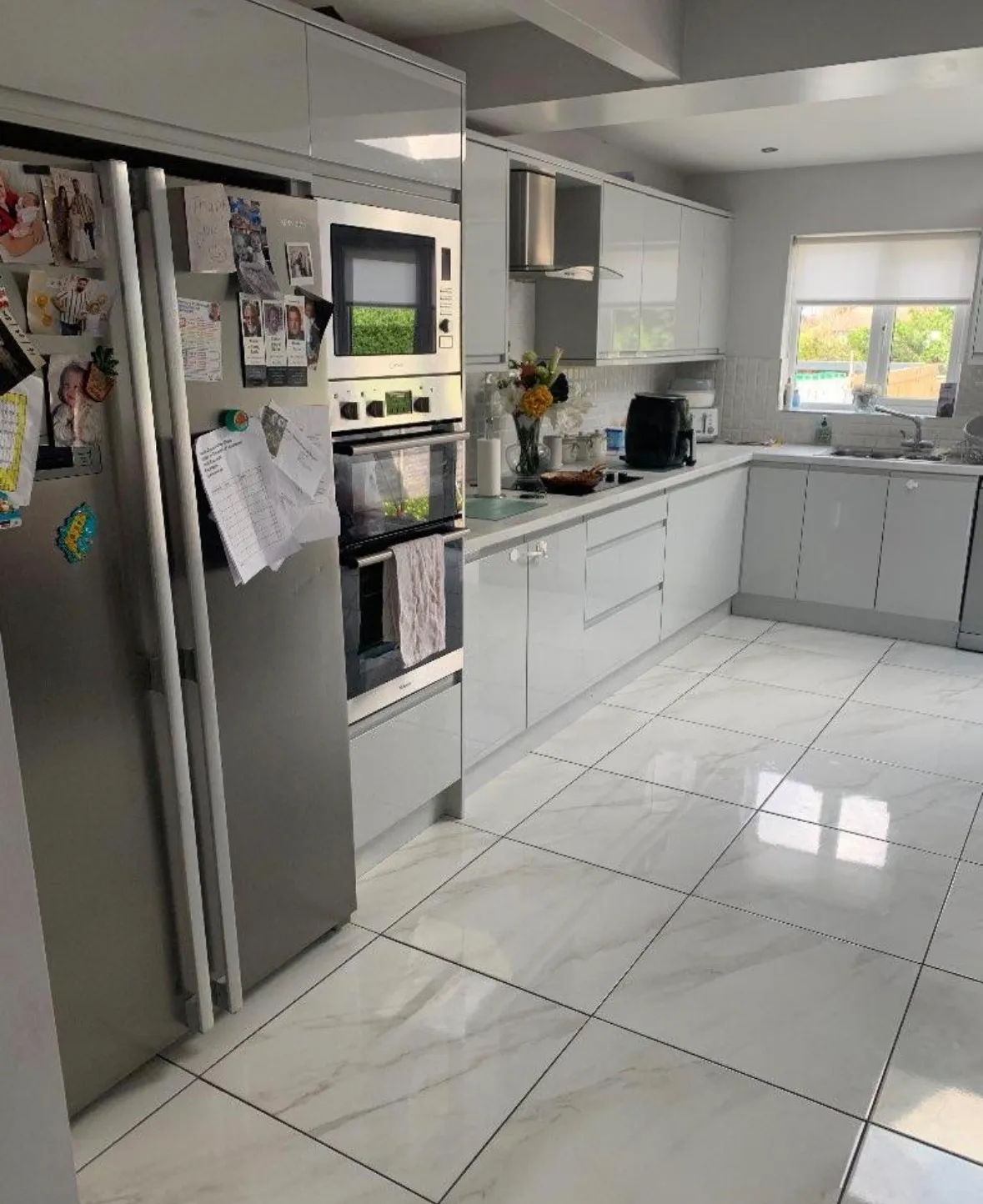 Kitchen with stainless steel refrigerator, double wall oven, L-shaped grey cabinets, sink under a window, countertop appliances, and glossy tiled floor.