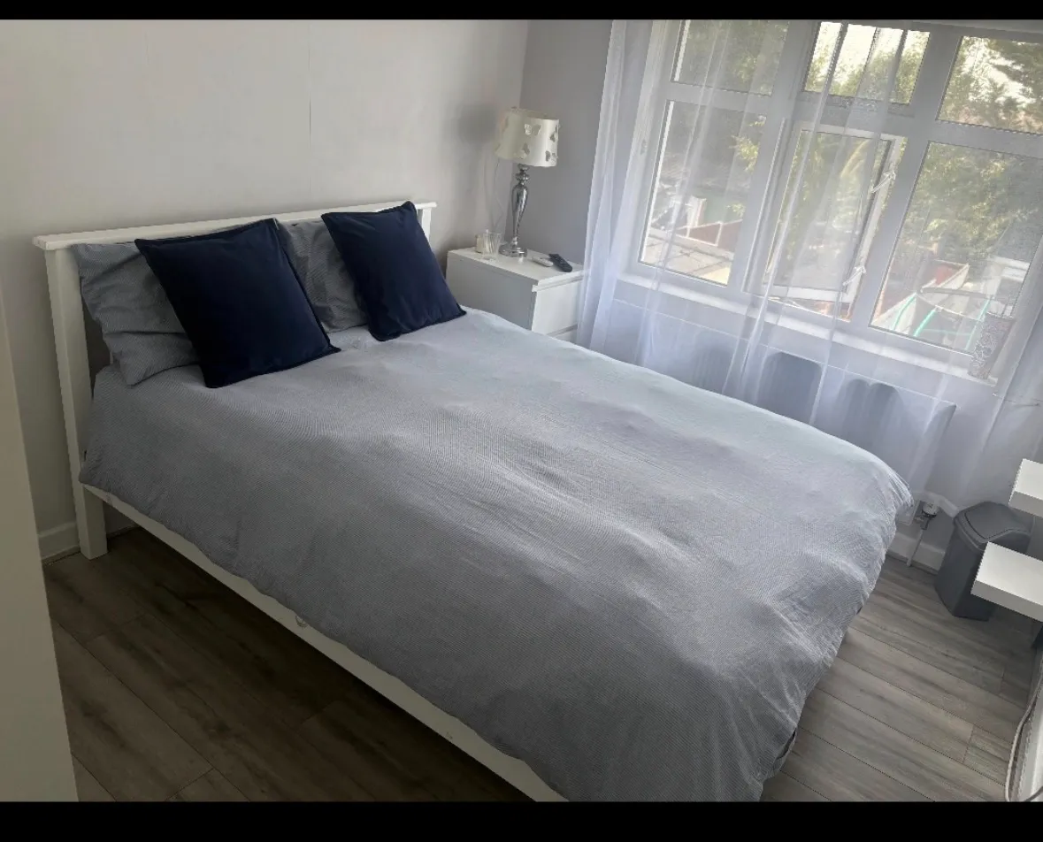Bedroom with a double bed made with light-gray bedding, two dark pillows, a nightstand with a lamp, and a large window with sheer curtains.