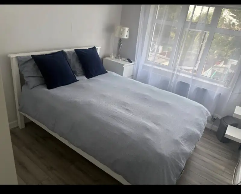 Bedroom with a double bed made with light-gray bedding, two dark pillows, a nightstand with a lamp, and a large window with sheer curtains.