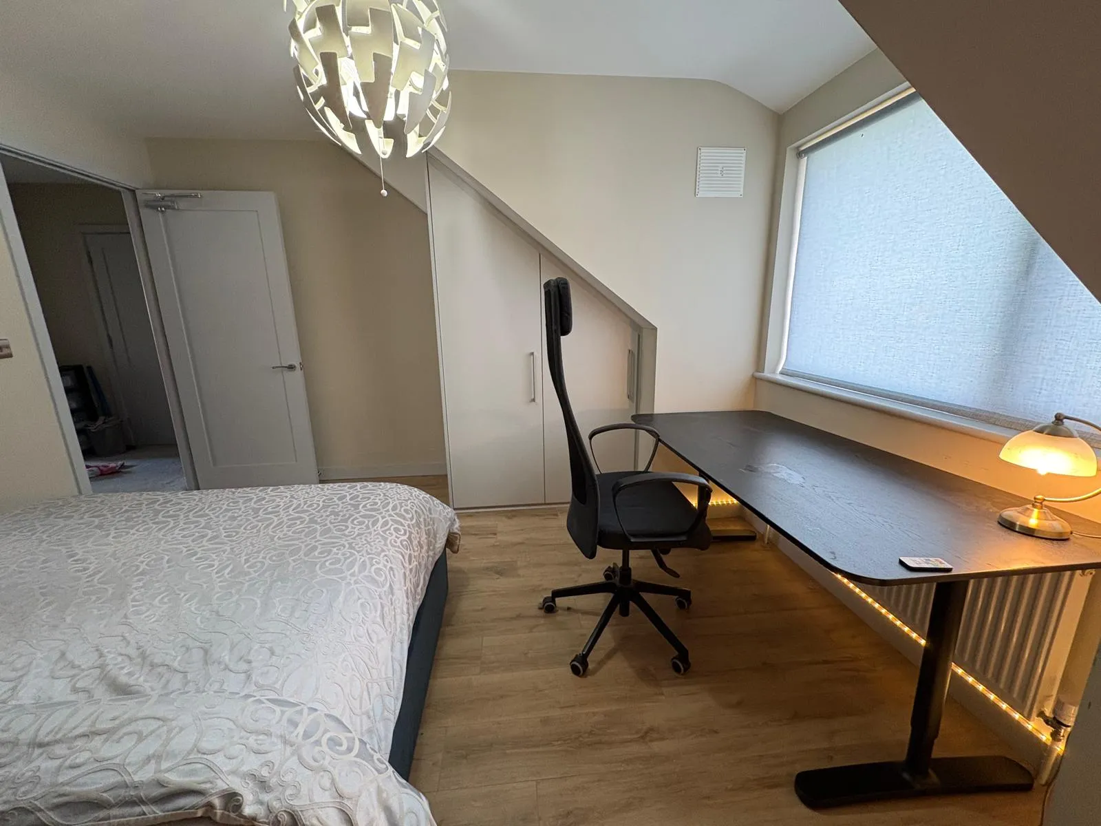 Bedroom with a bed, desk and office chair next to a window with a roller blind; built-in sloped wardrobe and pendant light.