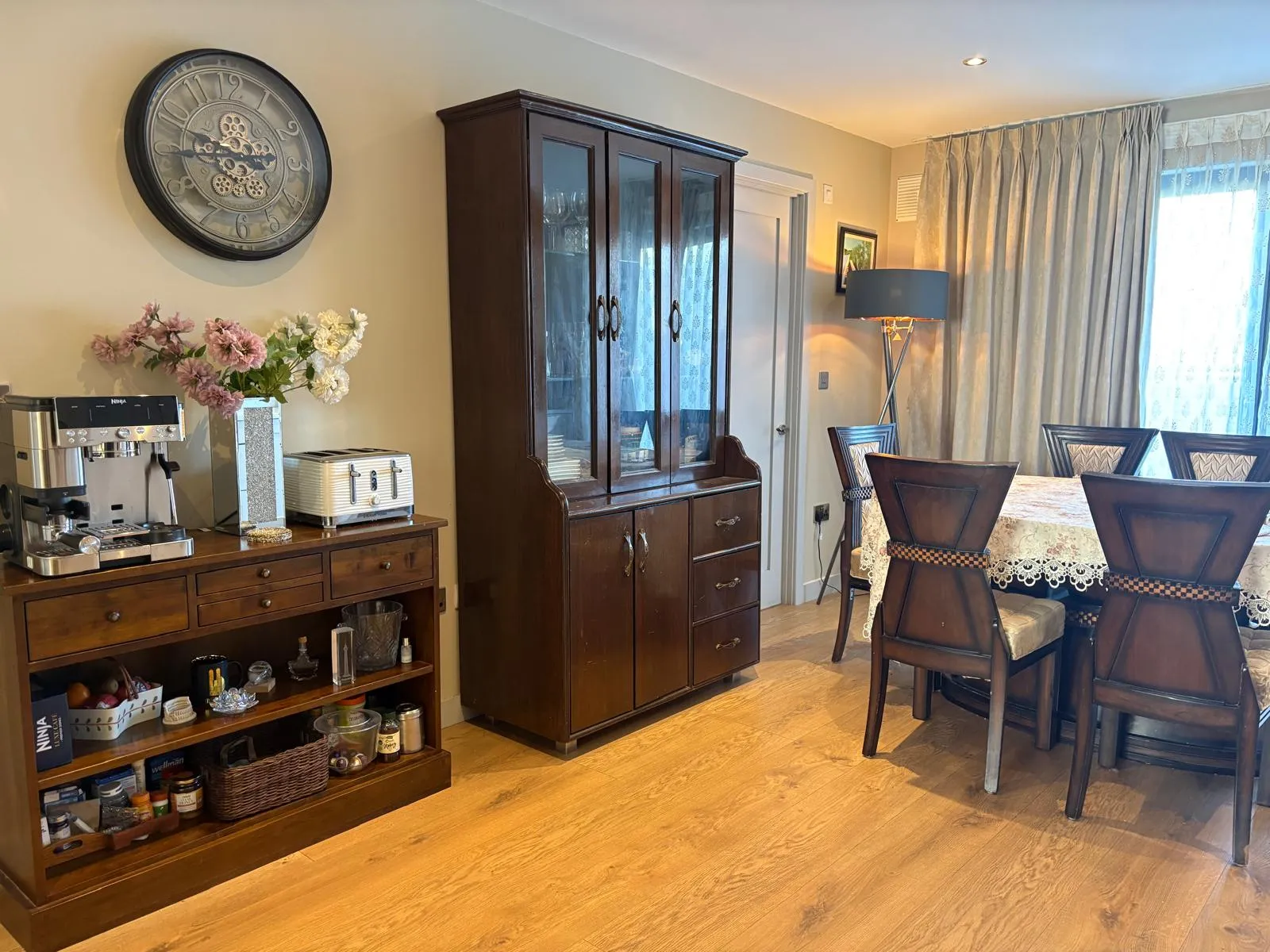 Dining room with a wooden dining table and chairs, tall glass-front cabinet, sideboard with coffee machine and toaster, wall clock and floor lamp near a curtained window.