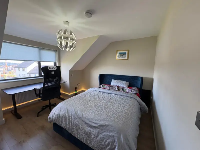 Bedroom with a double bed, upholstered headboard, desk and office chair by a large window, pendant ceiling light and sloped ceiling
