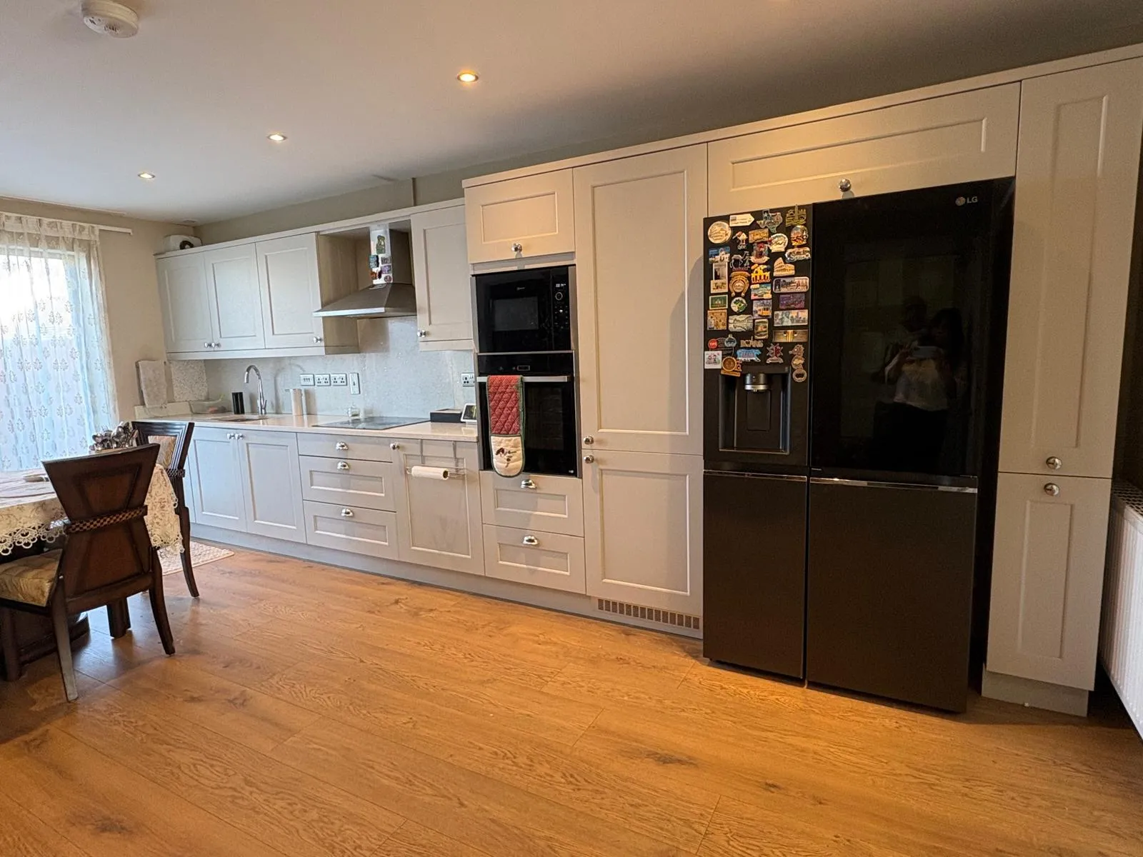 Kitchen with white cabinets, built-in oven, range hood, black refrigerator with magnets, sink and a small dining table at left.
