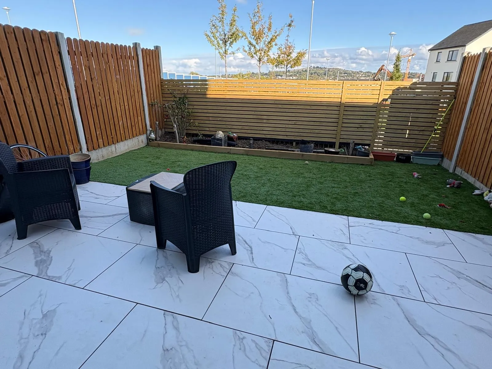 Small private backyard with tiled patio, two black outdoor chairs and a small table, artificial grass area, wooden privacy fence and a soccer ball on the patio.