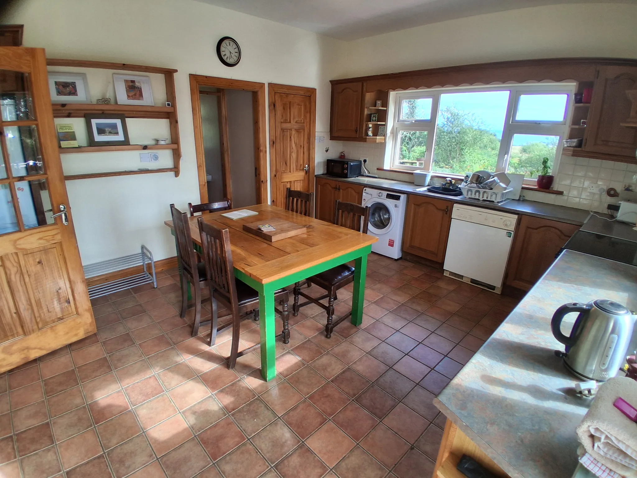 Kitchen with wooden cabinets, dining table and chairs, tiled floor, sink under a large window, washing machine and dishwasher.