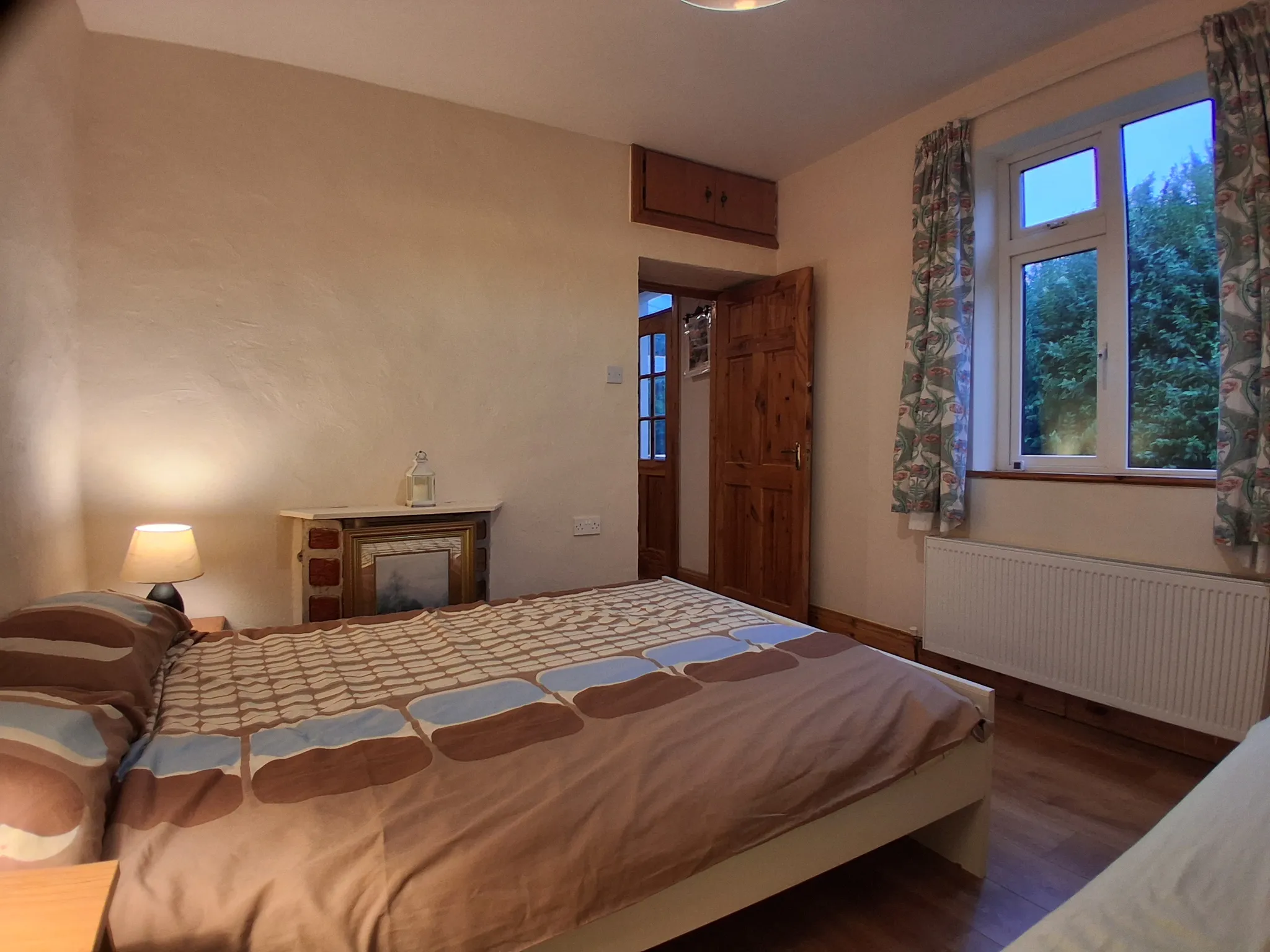 Bedroom with a double bed, bedside lamp and nightstand, wooden door, window with patterned curtains and a radiator.