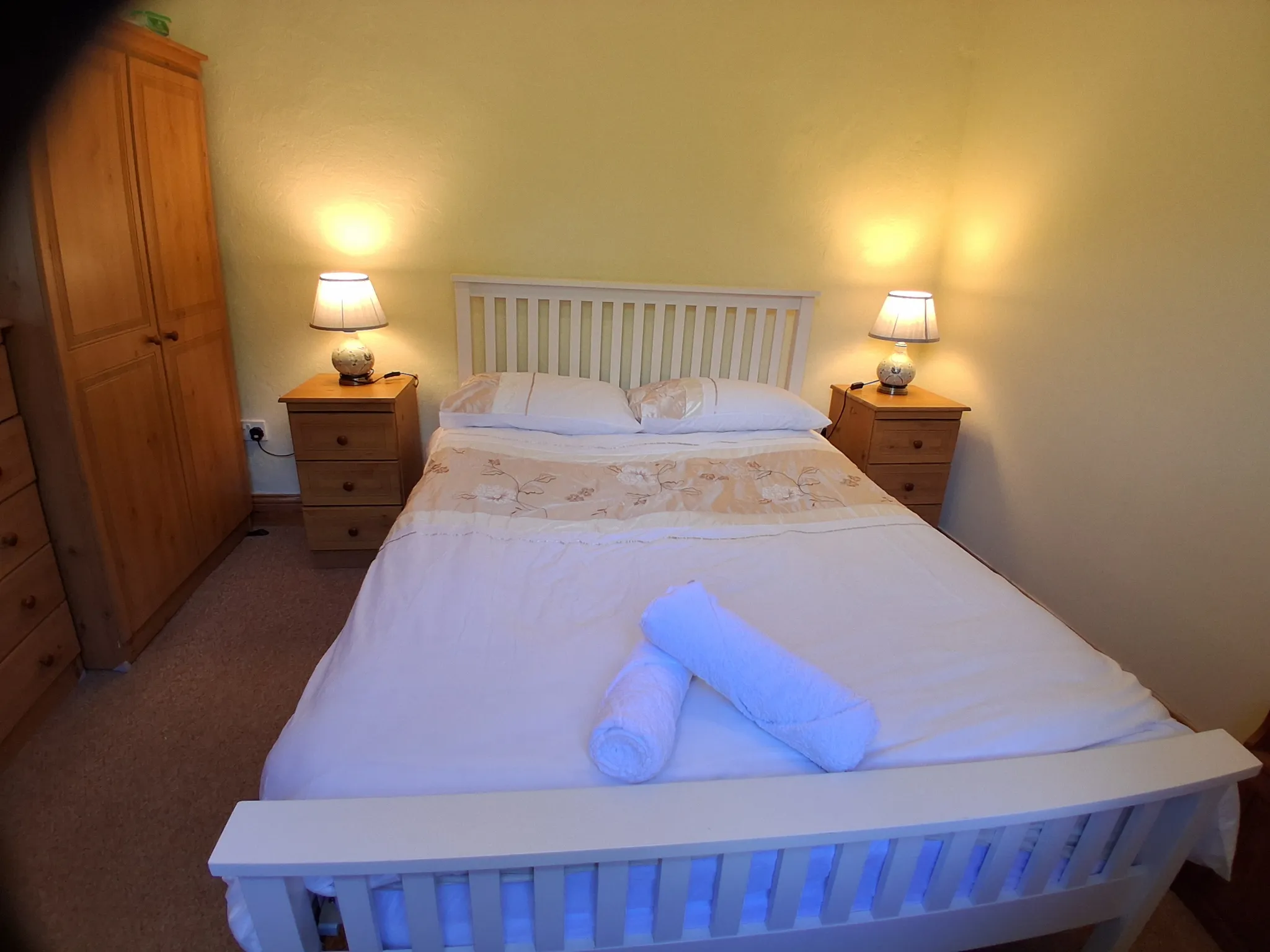 Bedroom with a double bed, two bedside tables with lamps, a wooden wardrobe to the left, and rolled towels on the bed.