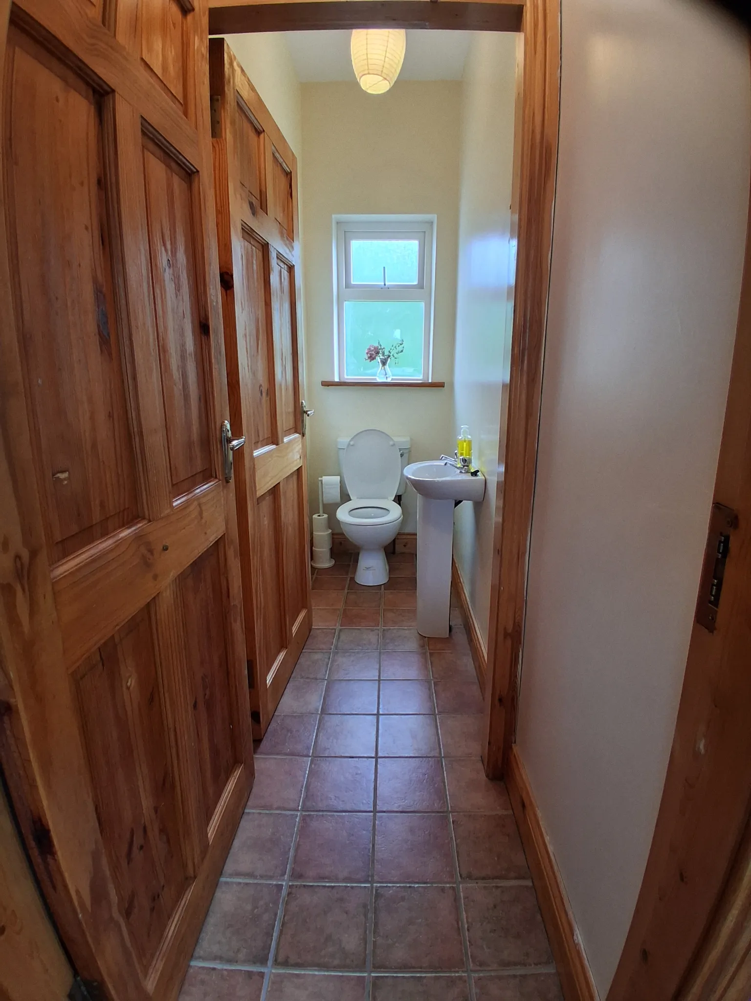 Small bathroom with a toilet, pedestal sink, small frosted window with a vase on the sill, pendant light and wooden doors.