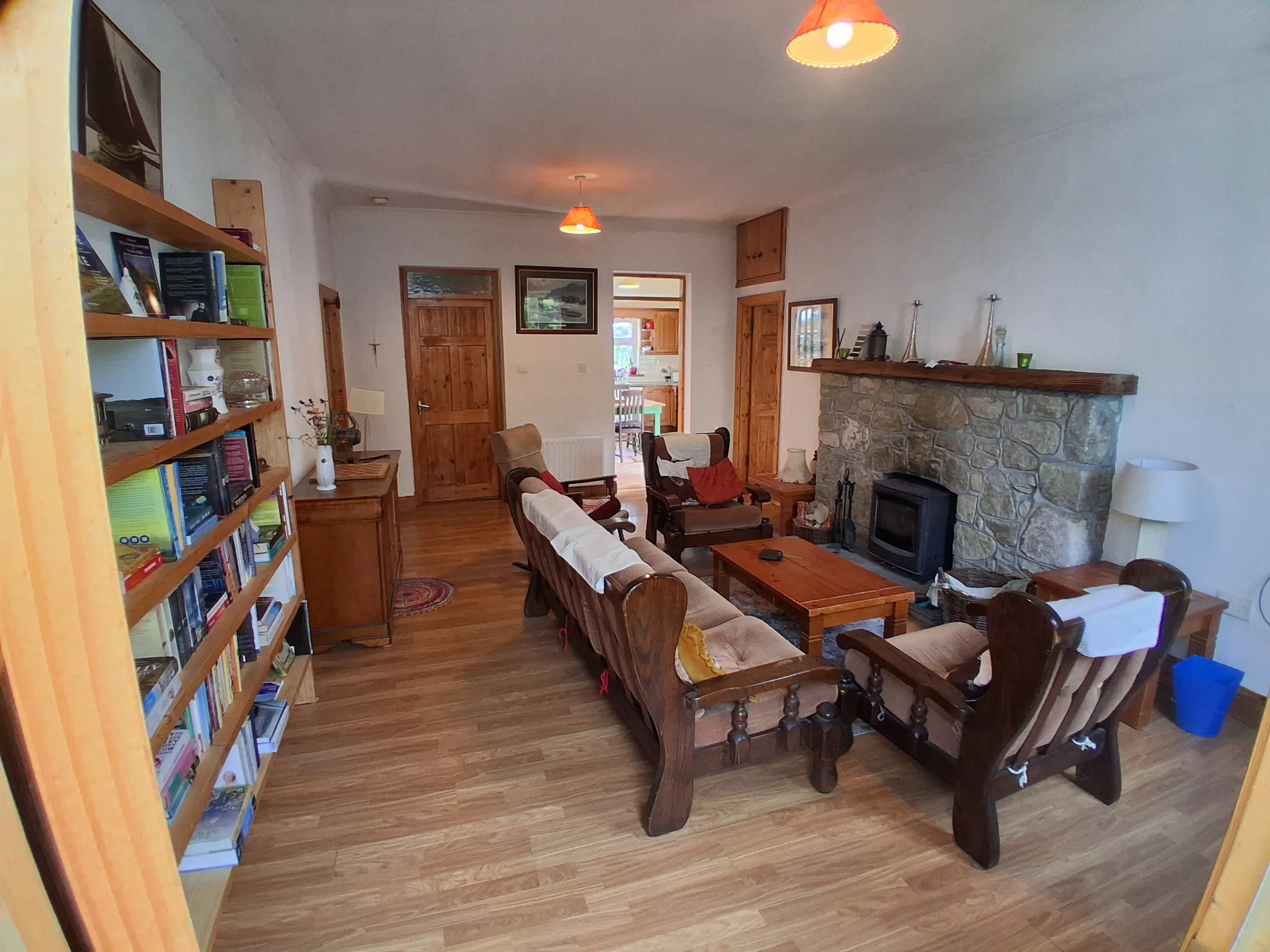 Living room with wooden sofas and armchairs around a wooden coffee table, stone fireplace with a wooden mantel, bookshelf on the left and a doorway leading to a kitchen.
