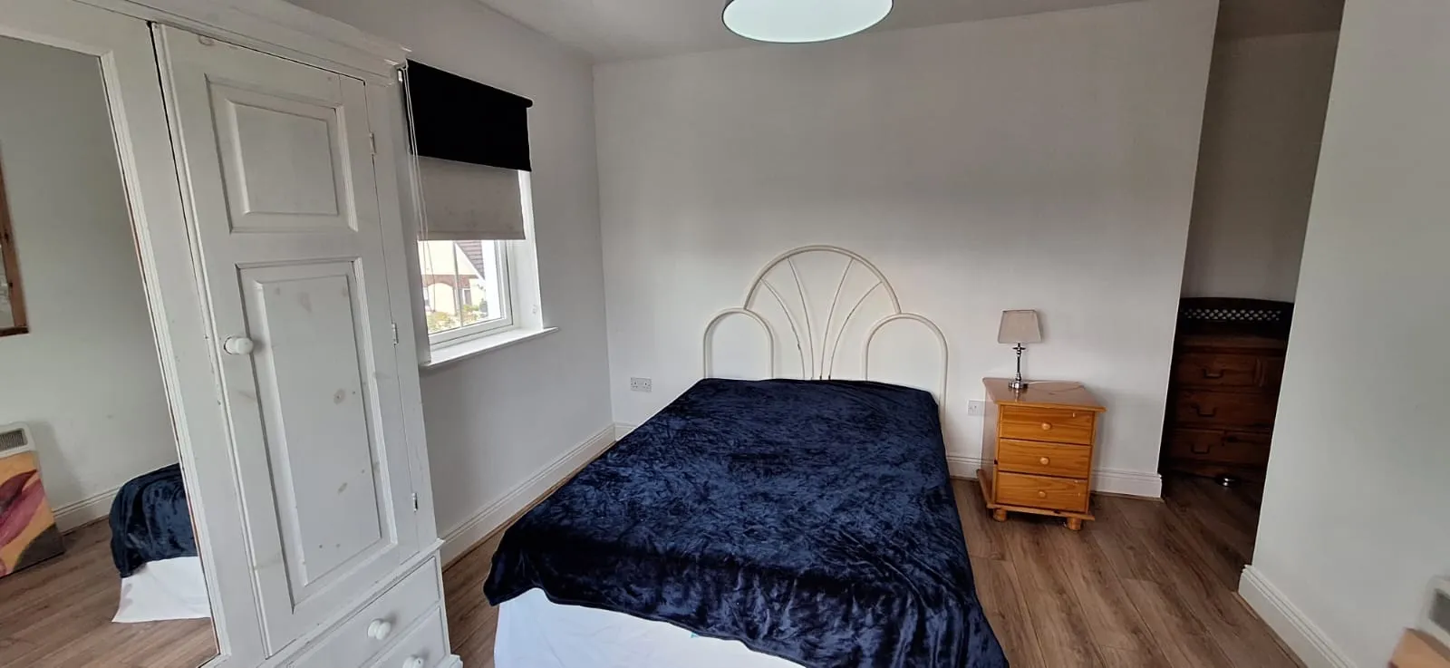 Bedroom with a double bed covered in a dark blanket, white metal headboard, wooden nightstand with a lamp, window with a roller blind, and a white wardrobe.