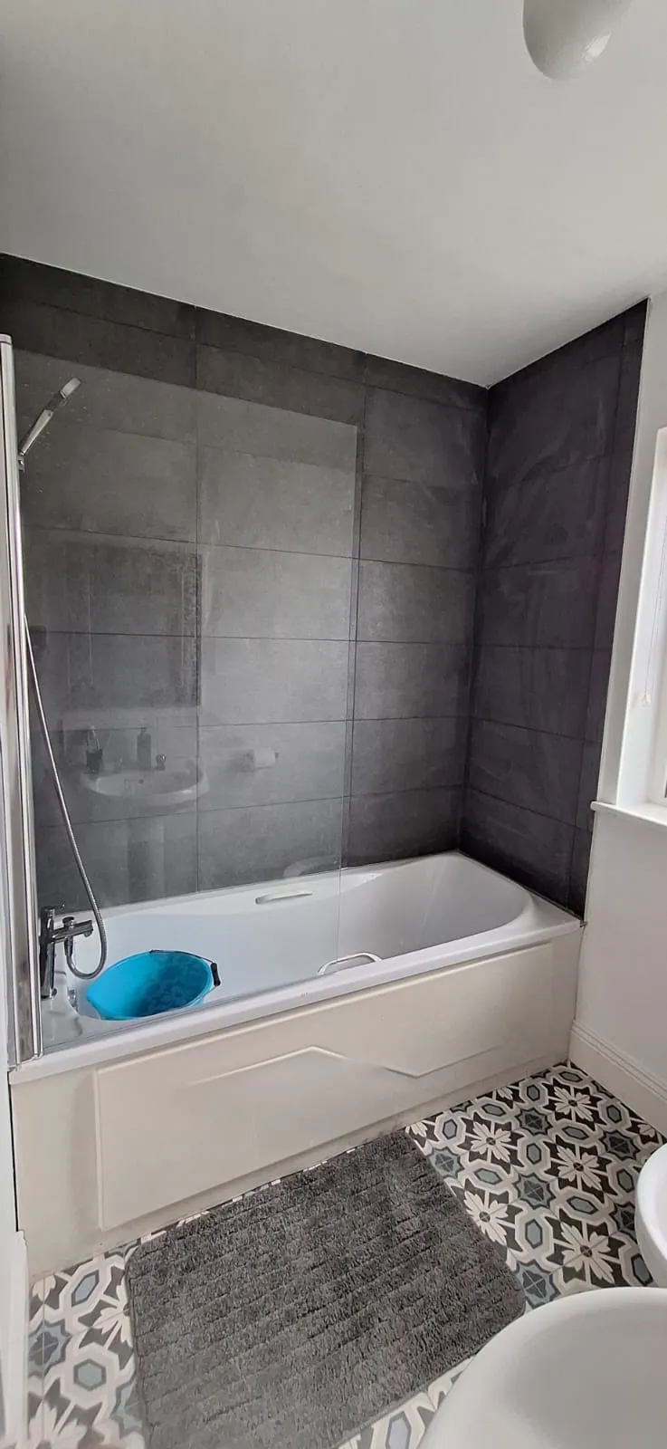 Bathroom with a bathtub, glass shower screen, dark tiled wall, patterned floor tiles, and a blue bucket in the tub.