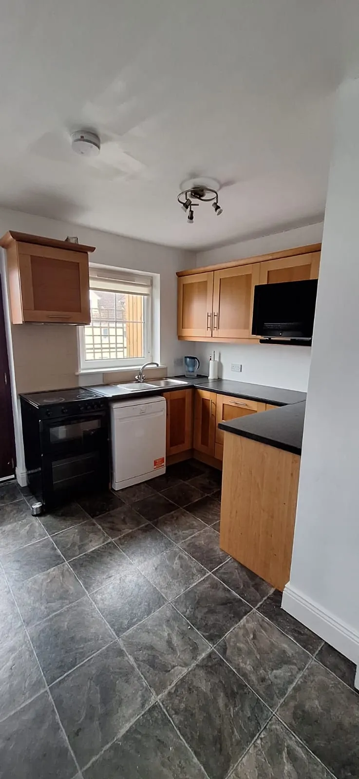 Kitchen with wood cabinets, black countertop, sink under a window, freestanding oven and dishwasher, and tiled floor.