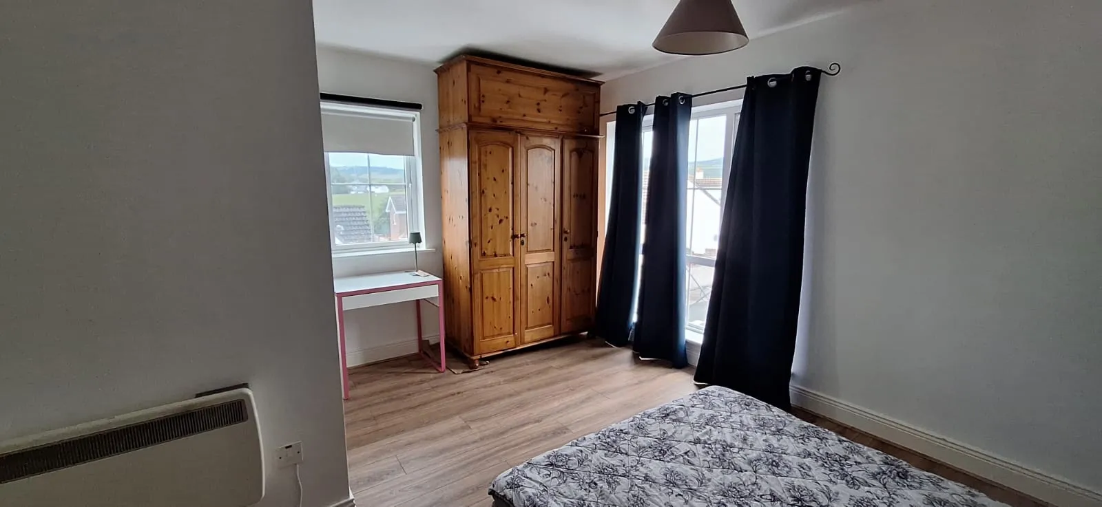 Bedroom with a double mattress on the floor, wooden wardrobe, small desk by a window with a blind, dark curtains on a larger window/door, and a wall-mounted heater.