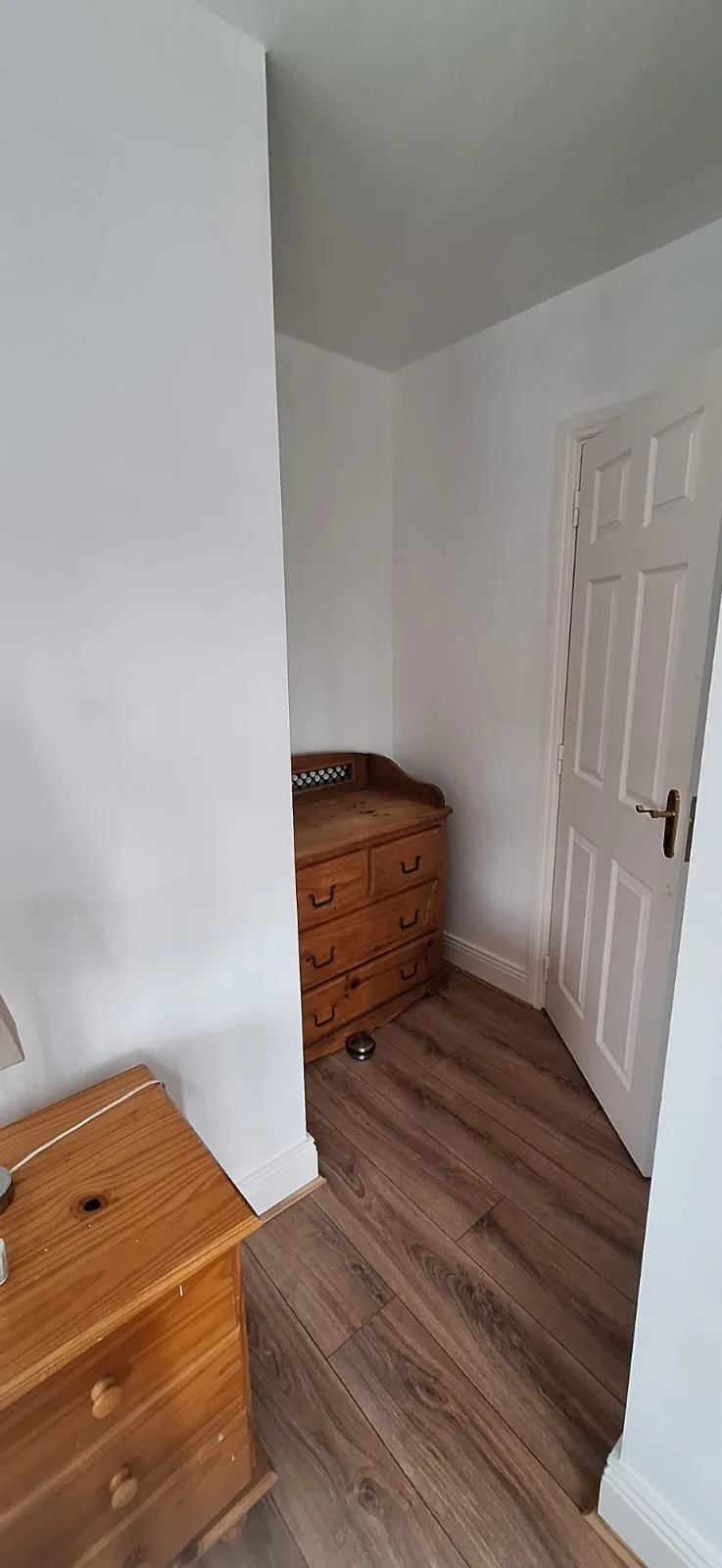 Bedroom corner with a wooden chest of drawers, a small bedside table, a white interior door, and wood-effect flooring.