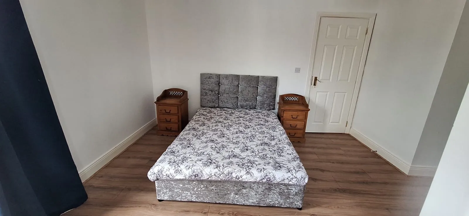 Bedroom with a double bed centered between two wooden bedside tables, upholstered headboard, closed white door on the right and dark curtains visible at left.