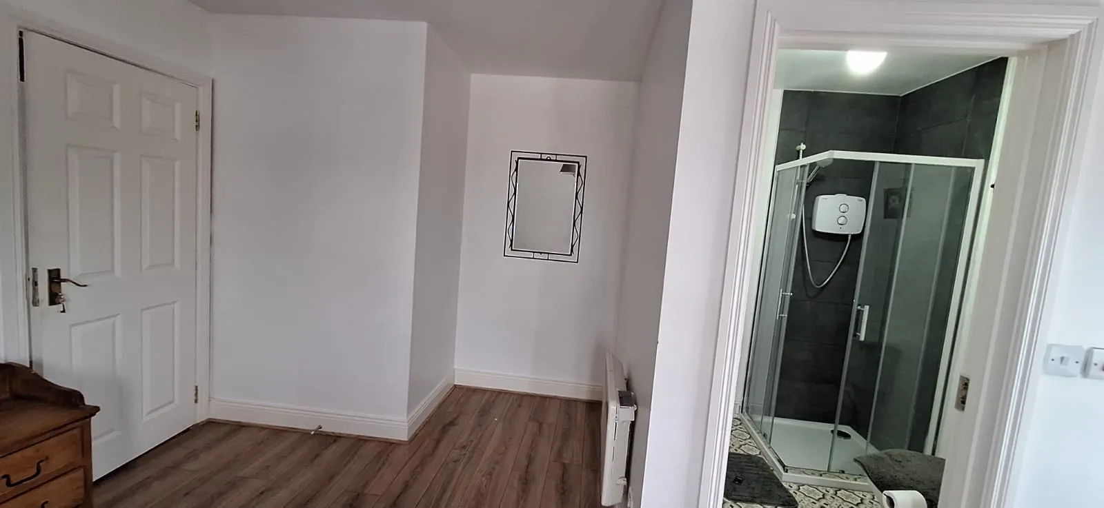 Room with wood-look floor, white walls, a closed white door, small wooden dresser at left, wall mirror, radiator, and an open doorway leading to a bathroom with a glass shower enclosure and electric shower unit.