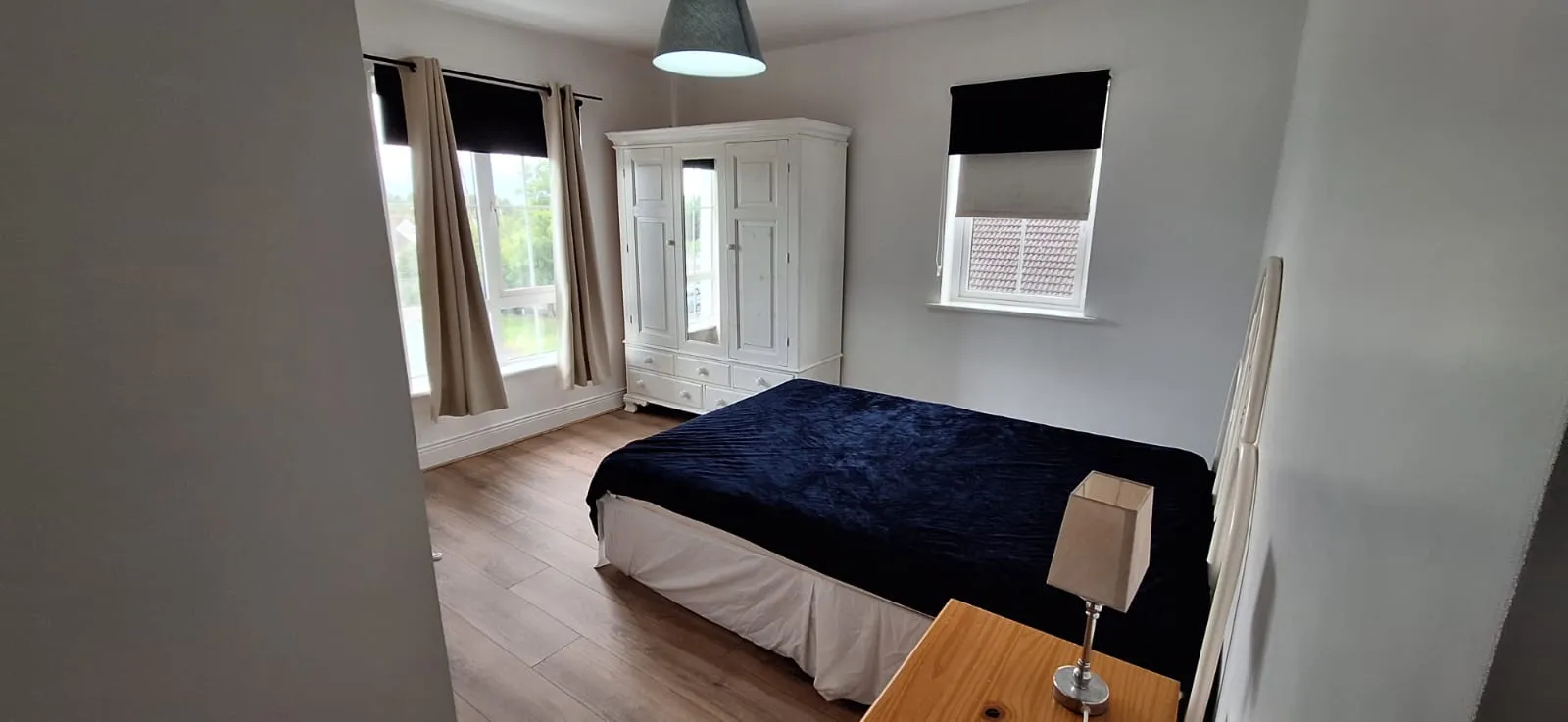 Bedroom with a double bed covered by a dark blanket, white wardrobe, two windows with curtains and blinds, wooden floor, and a bedside table with a lamp.