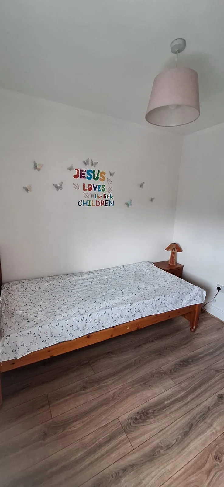 Bedroom with a single wooden bed, bedside table and lamp, wall decals reading "Jesus loves the little children", pendant ceiling light, and wood-look floor.