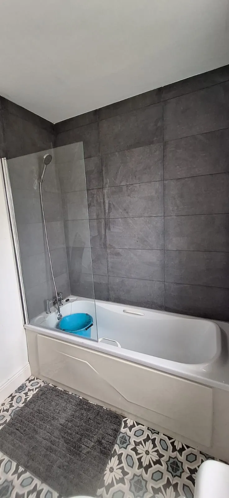 Bathroom with a bathtub and wall-mounted shower, glass shower screen, dark tiled wall and patterned floor with a grey bath mat and a blue bucket inside the tub.