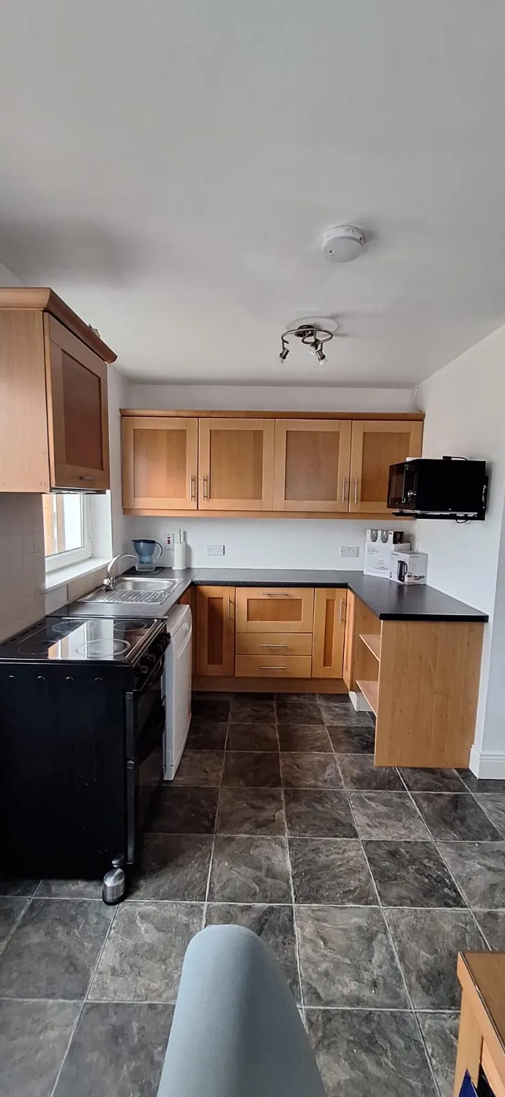 Kitchen with wooden upper and lower cabinets, black countertops, stainless sink under a window, freestanding stove, wall-mounted microwave, and dark tiled floor.