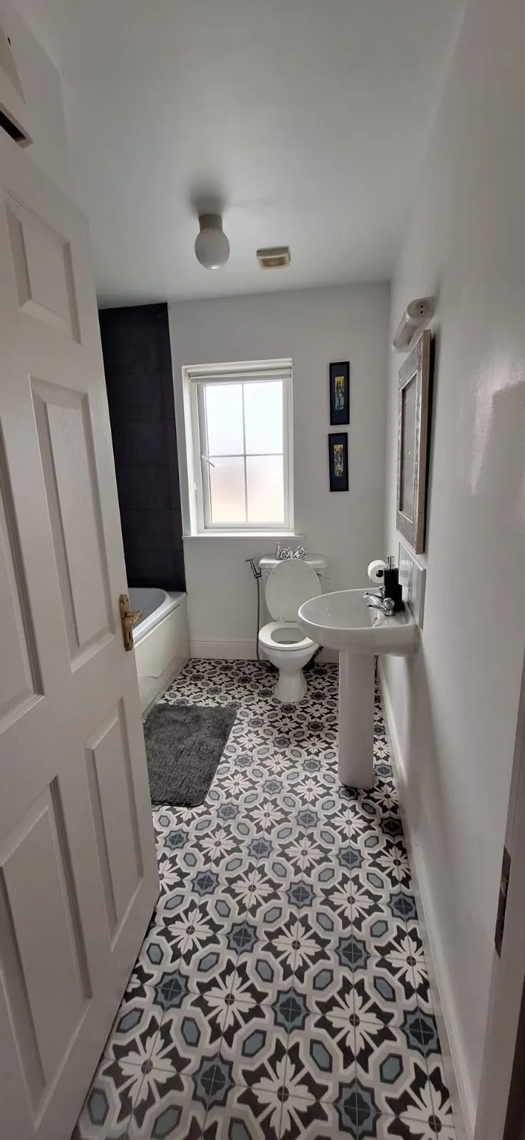 Bathroom with a pedestal sink, toilet, bathtub at left, patterned tile floor, window above the toilet, and a mirror on the right wall.