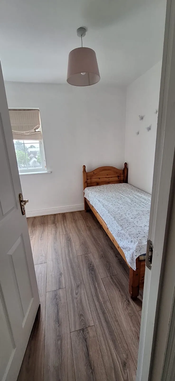 Small bedroom with a single wooden bed against the right wall, a window on the left, pendant ceiling light, and wood-look laminate floor.