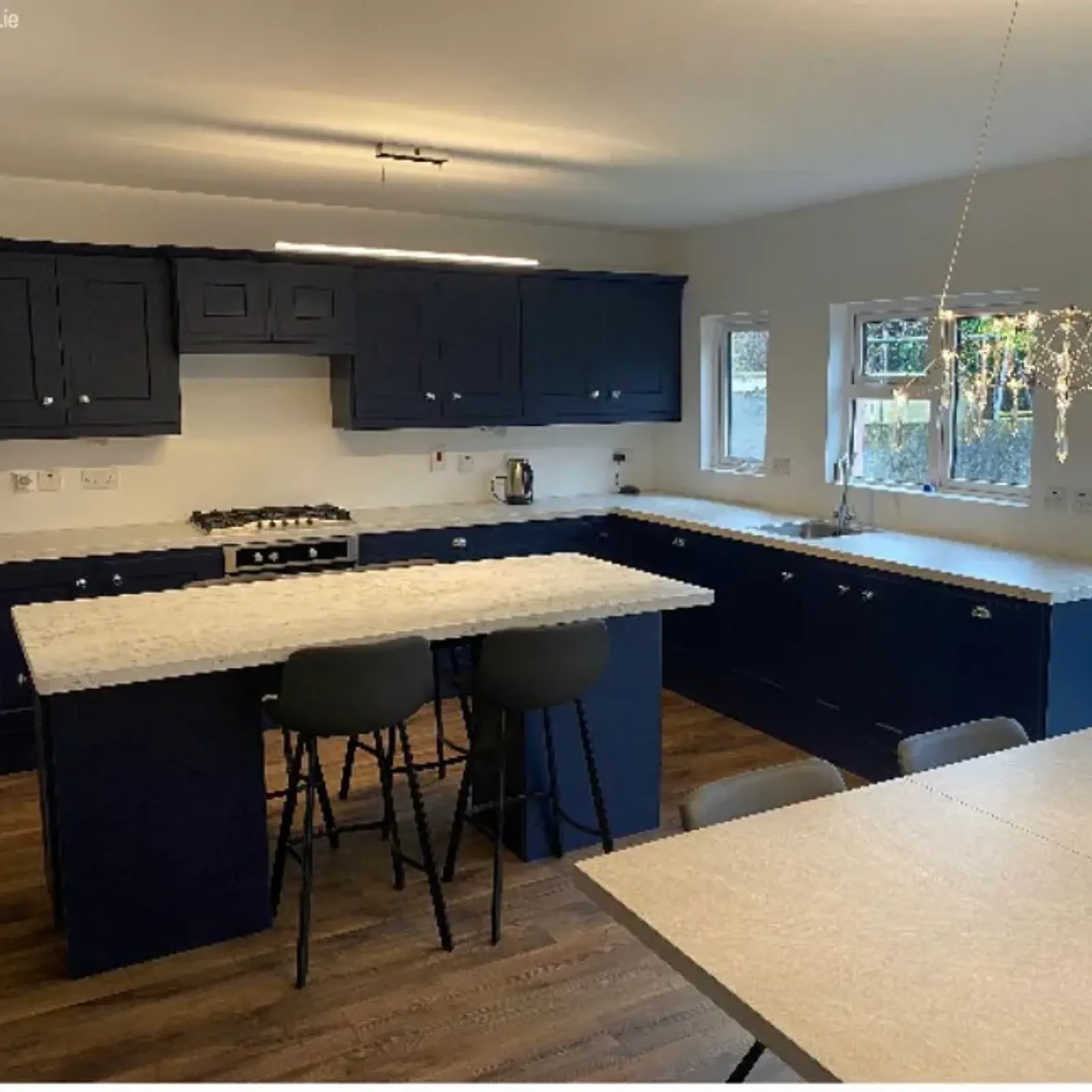 Kitchen with a central island, two bar stools, dark blue lower and upper cabinets, marble-effect countertops, gas stove, sink under two windows, and a hanging light fixture