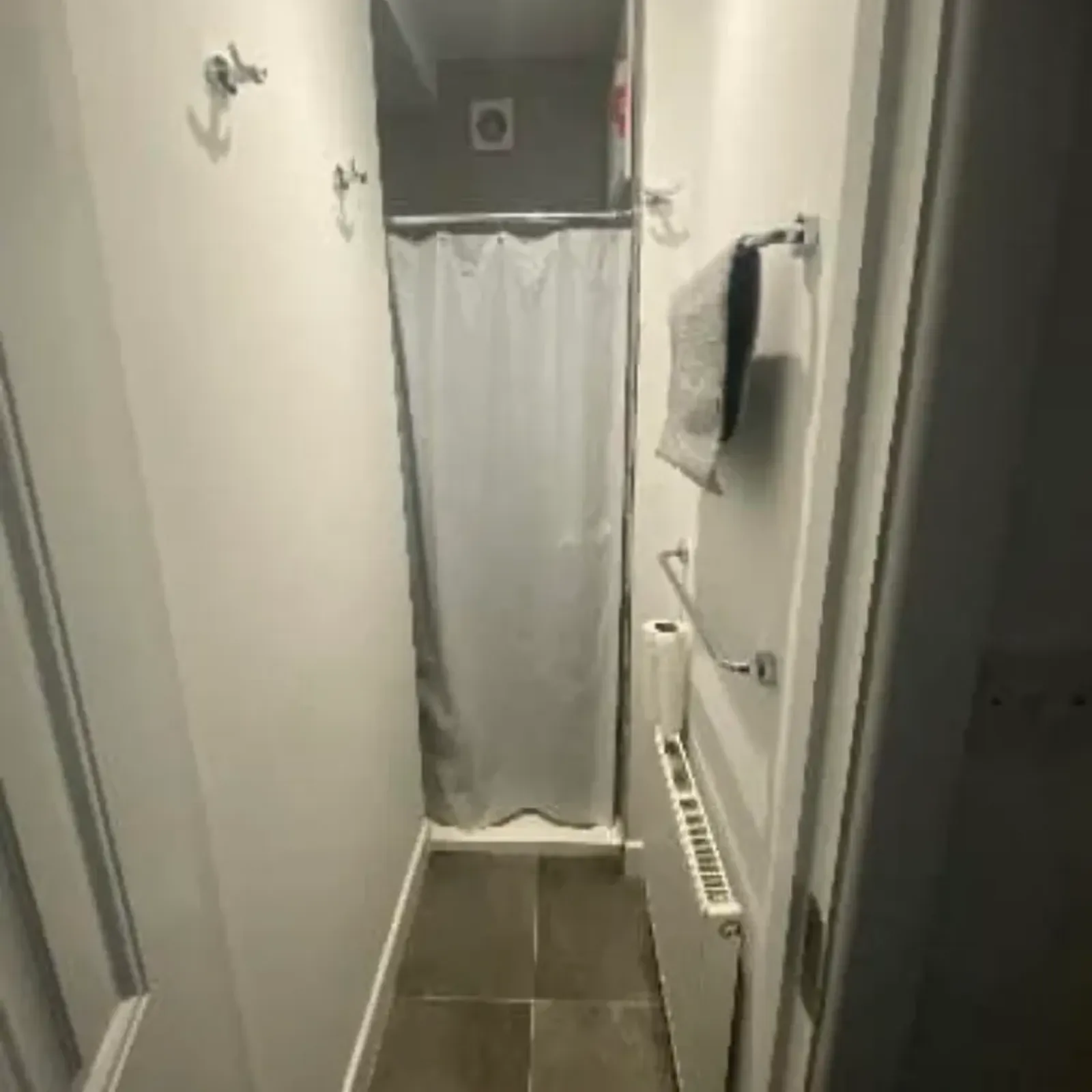 Narrow bathroom corridor with tiled floor, radiator and towel rack on the right, wall hooks on the left, and a shower with a closed curtain at the far end.