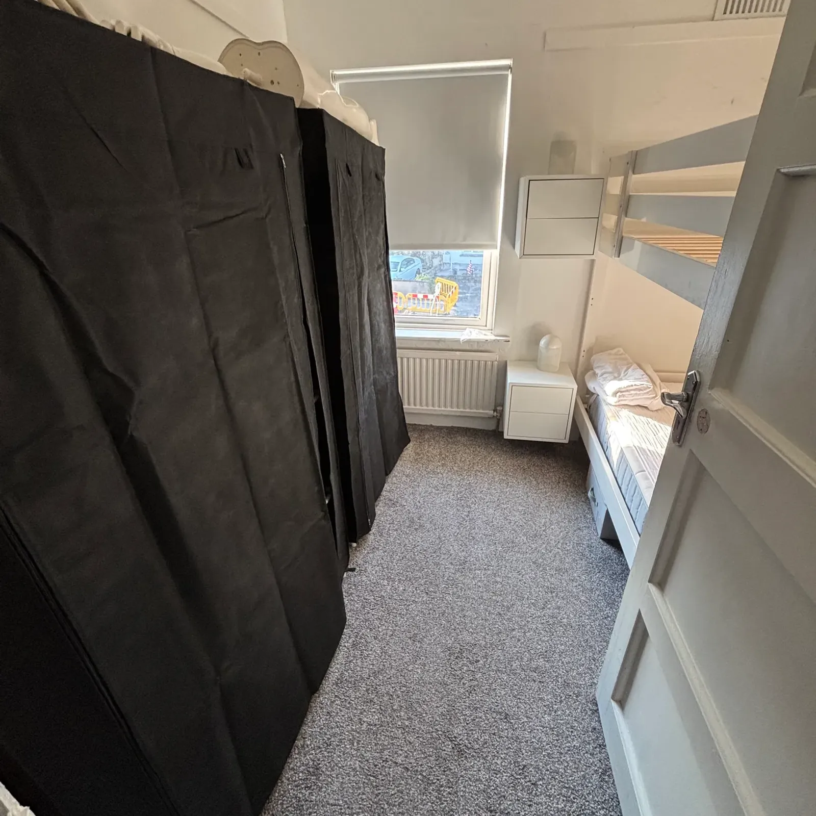 Small bedroom with a bunk bed on the right, fabric-covered wardrobe on the left, a window with a roller blind, small bedside table and radiator.