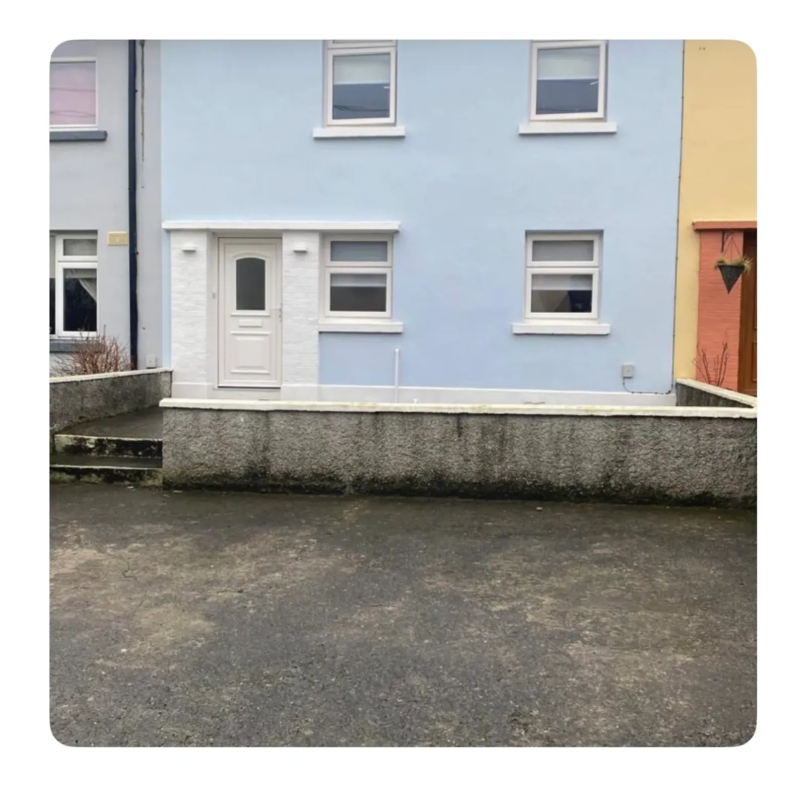 Front exterior of a two-story pale blue house with a white entrance door, three ground and first-floor windows, a low concrete wall and paved area in front.