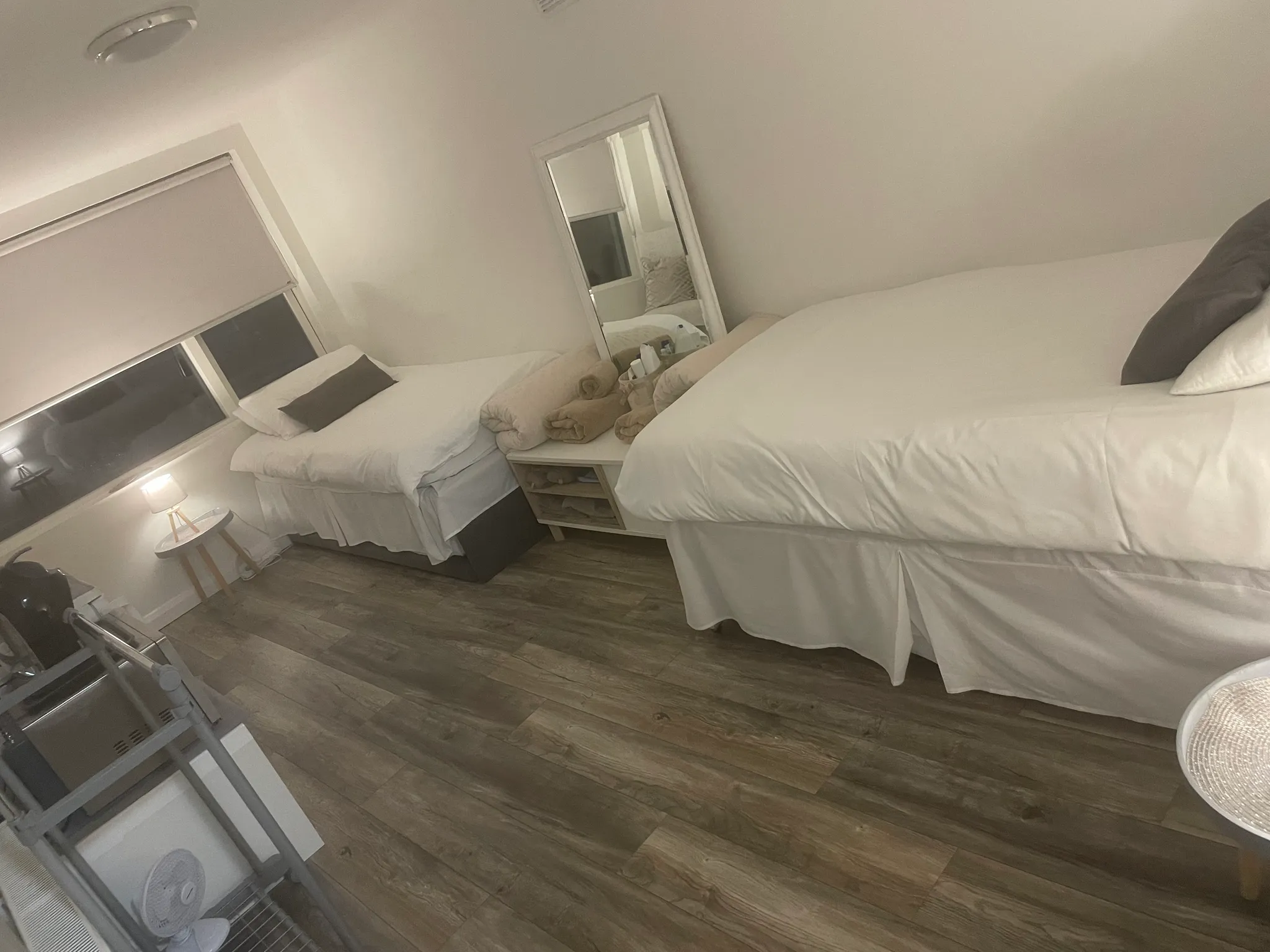 Bedroom with two single beds, a full-length mirror between them, a bedside table with rolled towels, a small lamp and a window with a blind