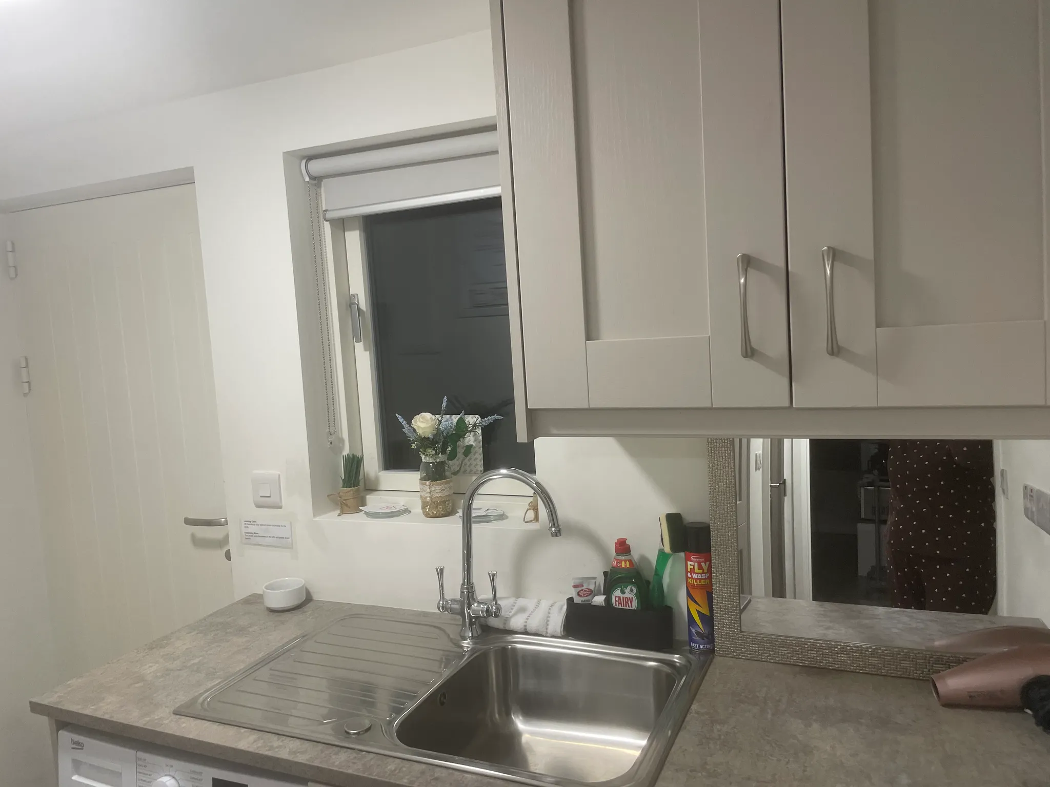Kitchen sink and countertop with a single-bowl stainless steel sink, chrome faucet, upper cabinets, a small window with a roller blind, and cleaning products on the counter.