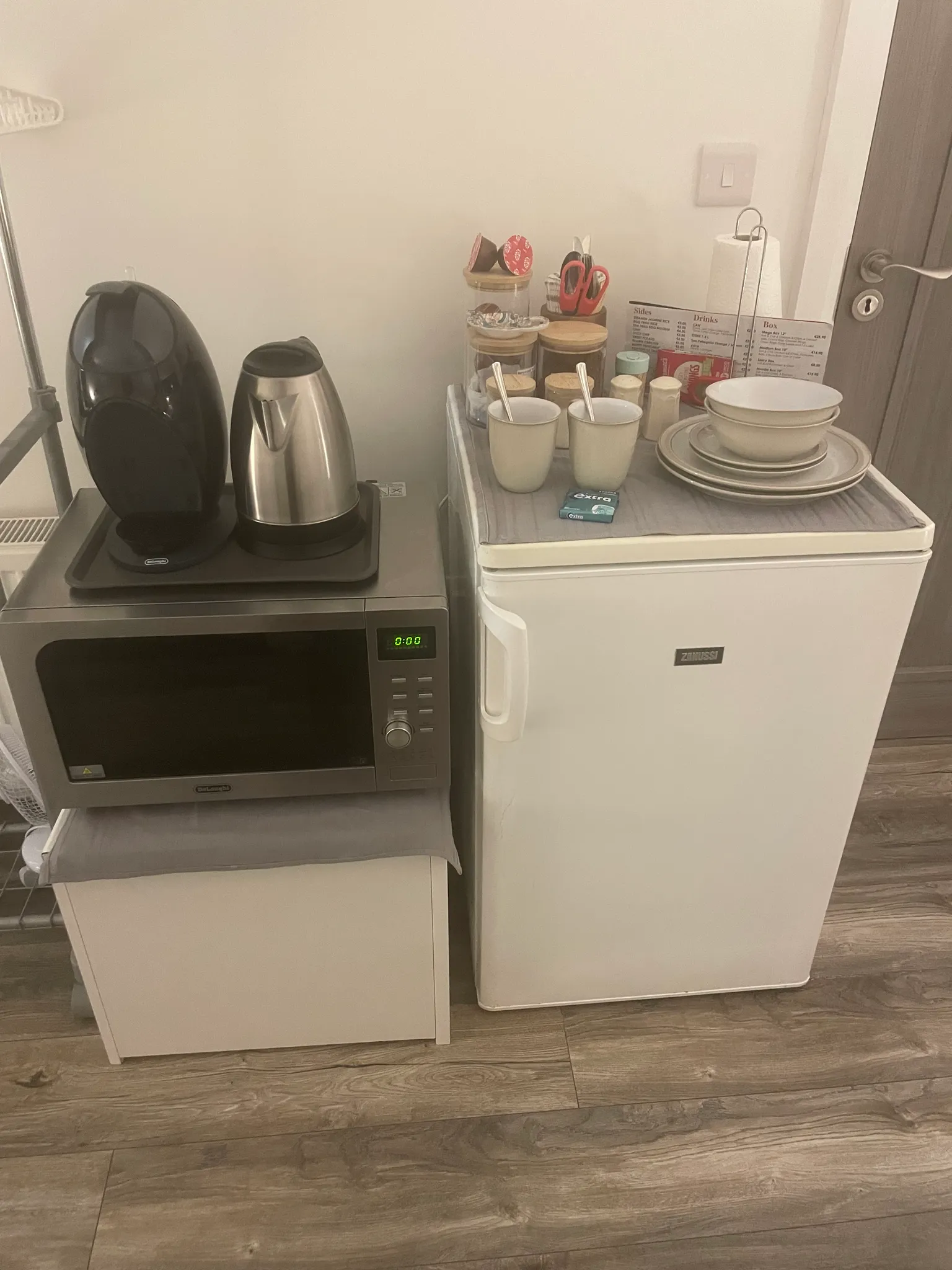 Small kitchenette counter with a microwave, electric kettle, mini fridge and stacked dishes with mugs and spoons on top