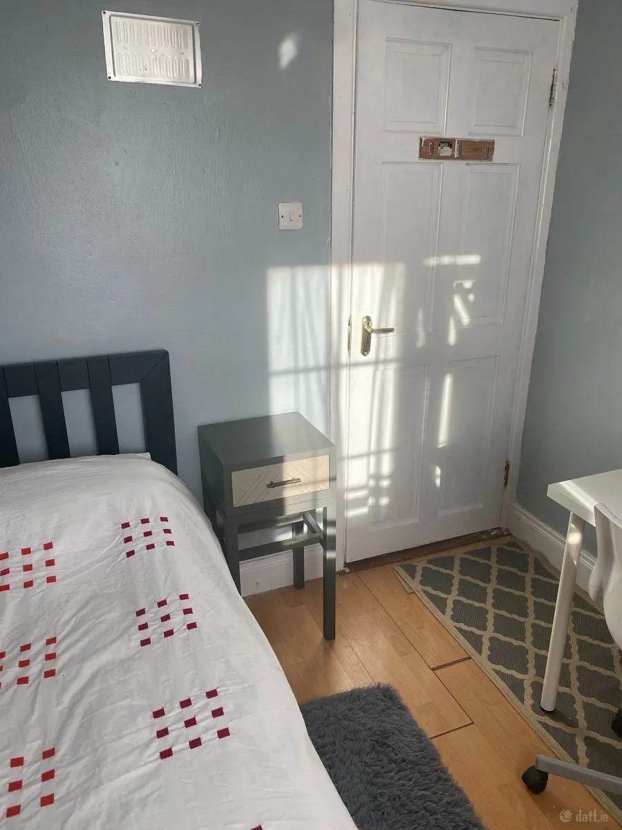 Bedroom with a single bed and patterned white duvet, a small bedside table, white paneled door, desk chair and rug; sunlight casting shadows on the door.