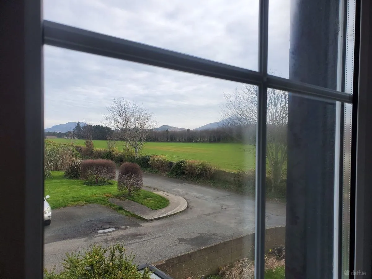 View through a window showing a driveway and lawn in the foreground, shrubs and hedges, open green fields and distant low hills under an overcast sky.