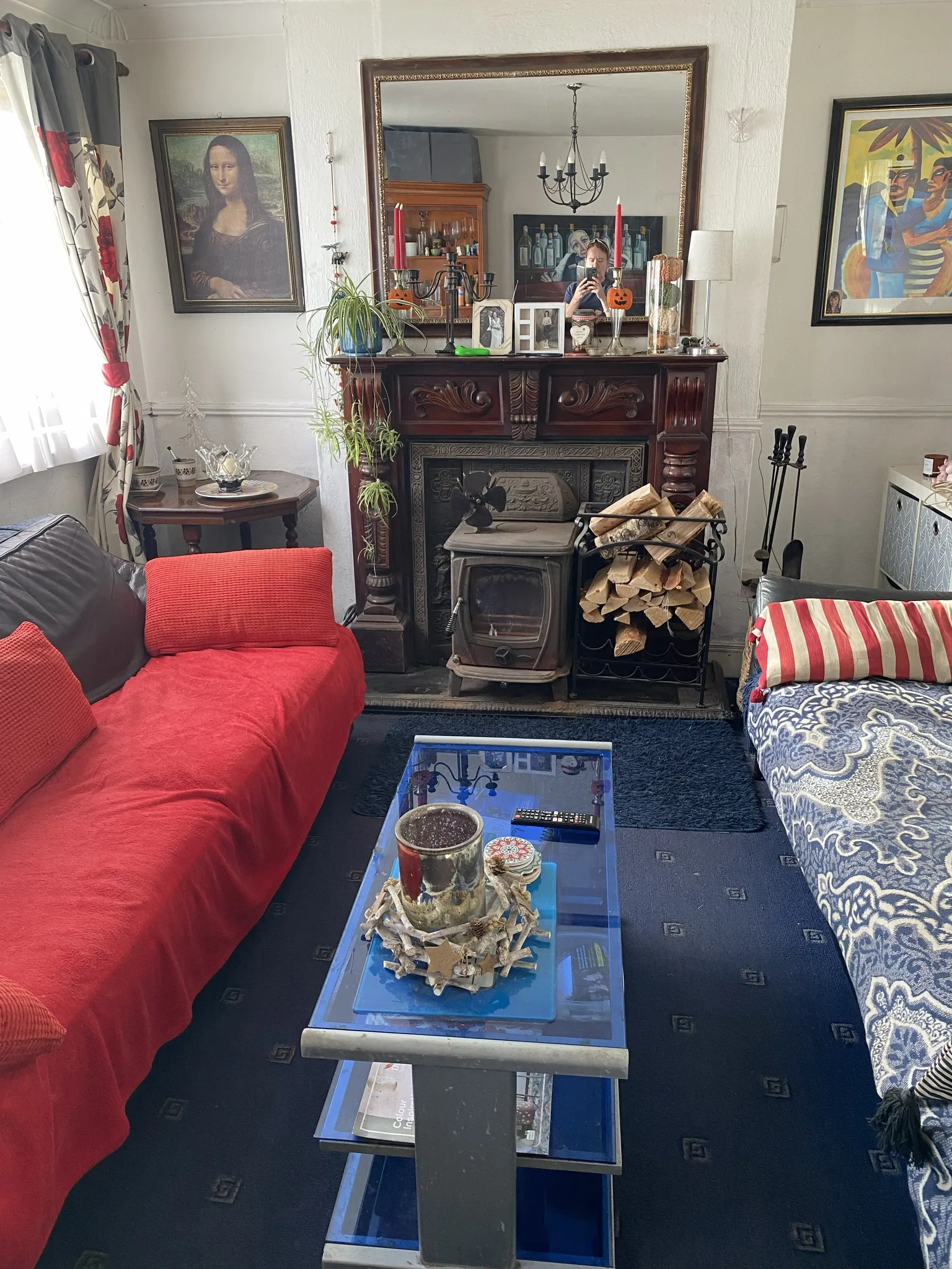 Living room with a wood-burning stove set into a decorative wooden mantel, a mirror above, red-covered sofa on the left, patterned sofa on the right, and a glass coffee table in the center.