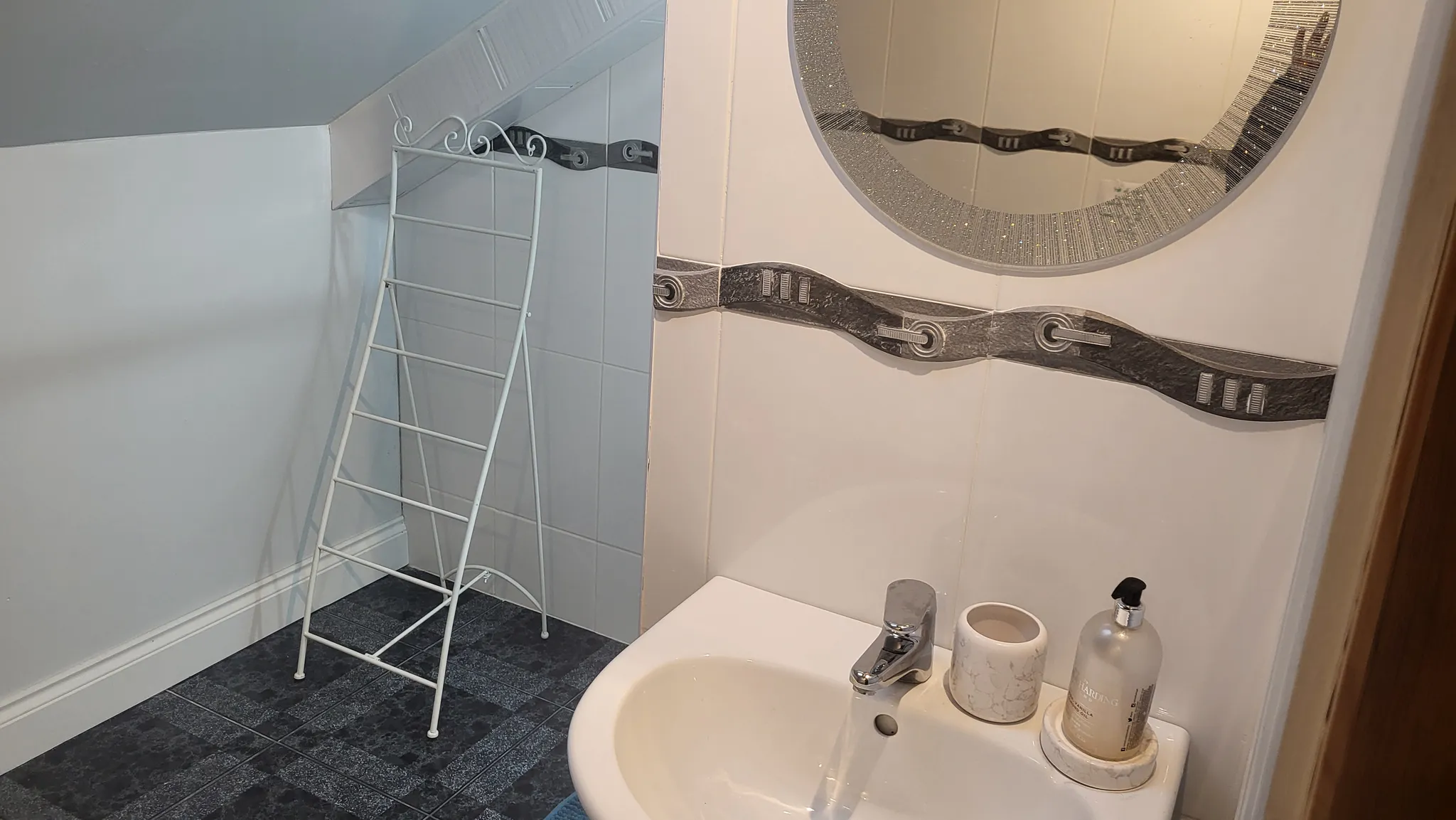 Bathroom sink with faucet, soap dispenser and cup, round wall mirror, and a white metal drying rack under a sloped ceiling