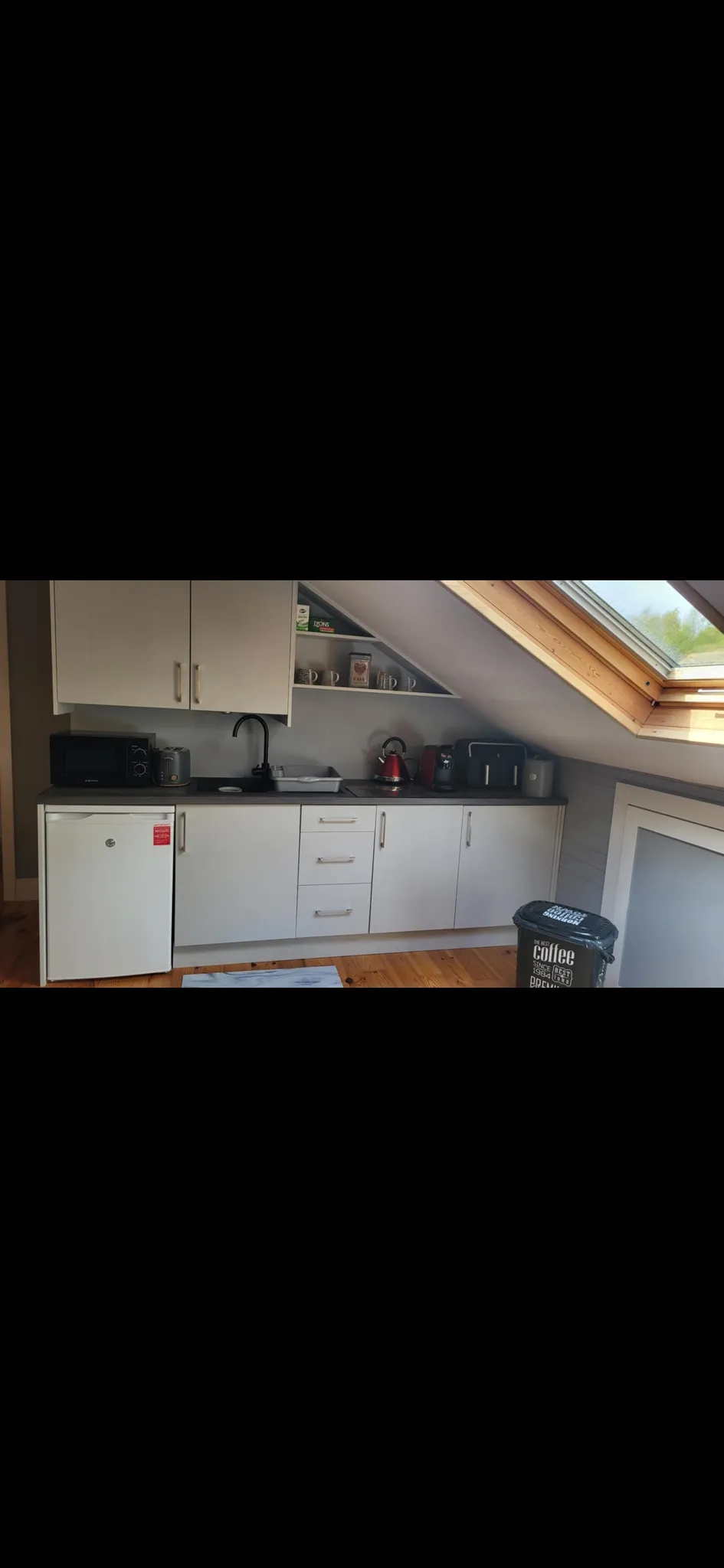 Small kitchen area under a sloped ceiling with white base and wall cabinets, countertop with sink and faucet, mini fridge, microwave, kettle, toaster and a skylight window above.