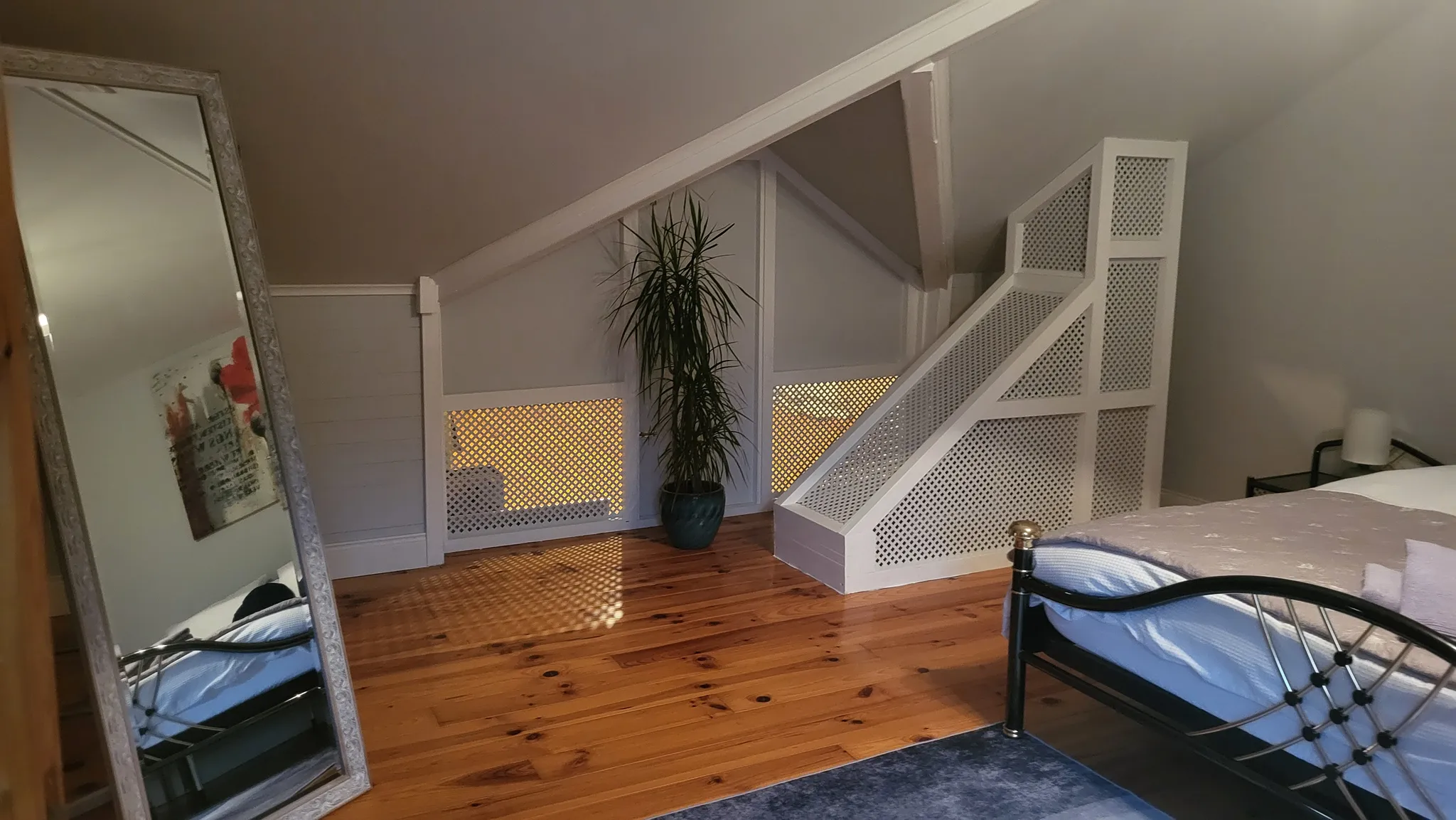 Bedroom under a sloped ceiling with a metal bed frame, hardwood floor, full-length mirror, potted plant and white lattice radiator covers
