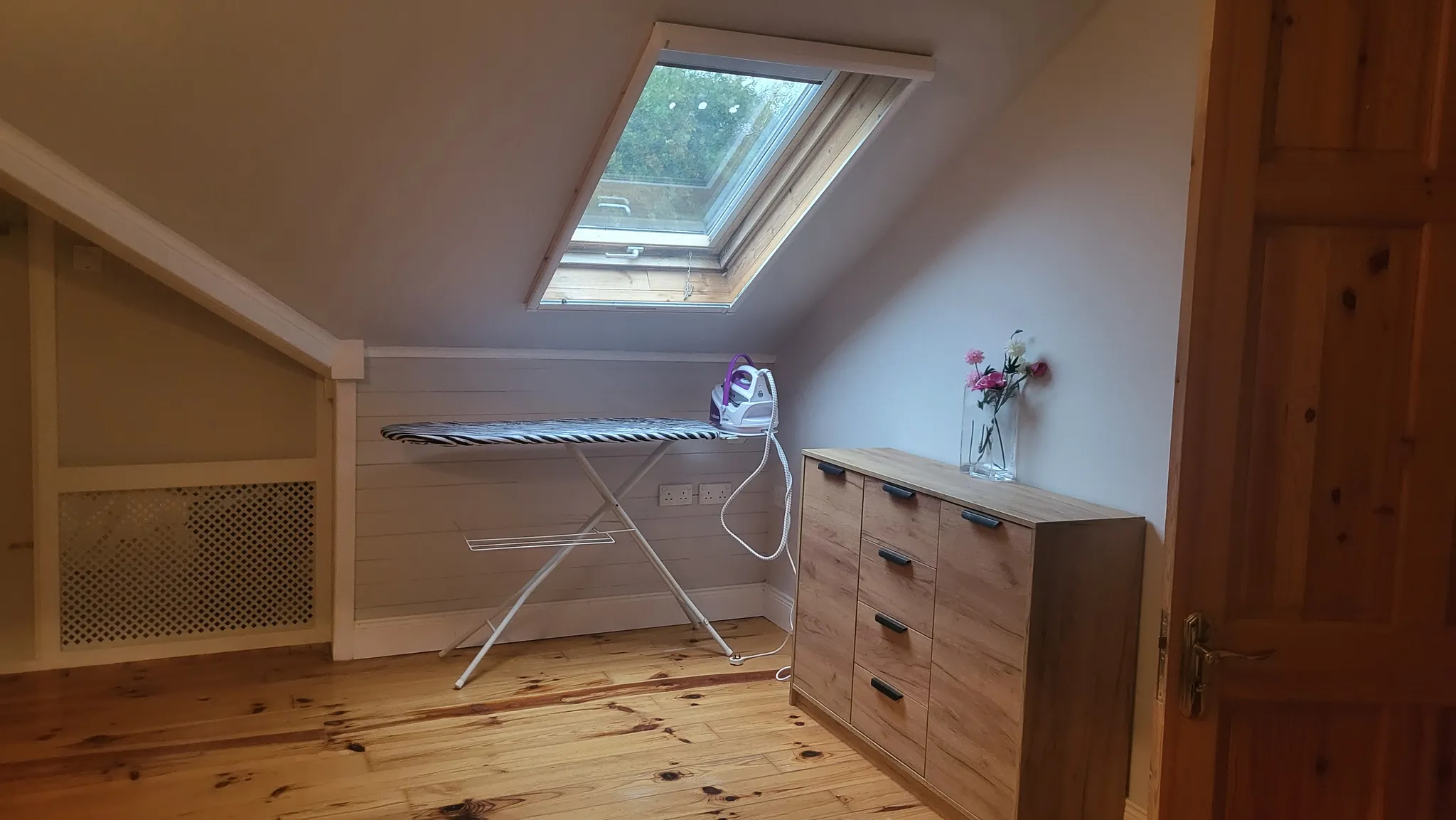 Attic room with a sloped ceiling and skylight, an ironing board with an iron, and a wooden chest of drawers with a vase of flowers.